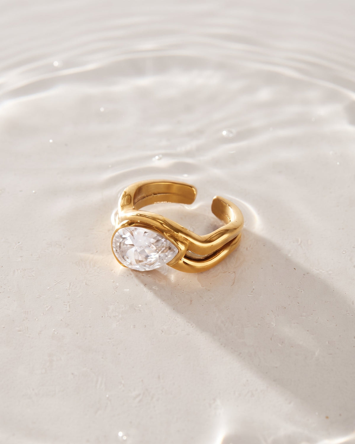 A gold adjustable ring with a teardrop-shaped clear gemstone, resting on a light beige surface with water ripples in the background.
