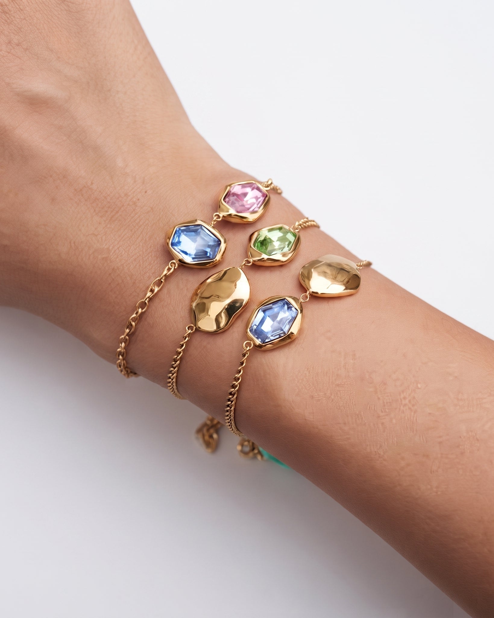 A close-up of a person's wrist wearing three layered gold bracelets, each decorated with a different faceted gemstone in pink, green, and blue, alongside textured gold leaf-shaped charms.