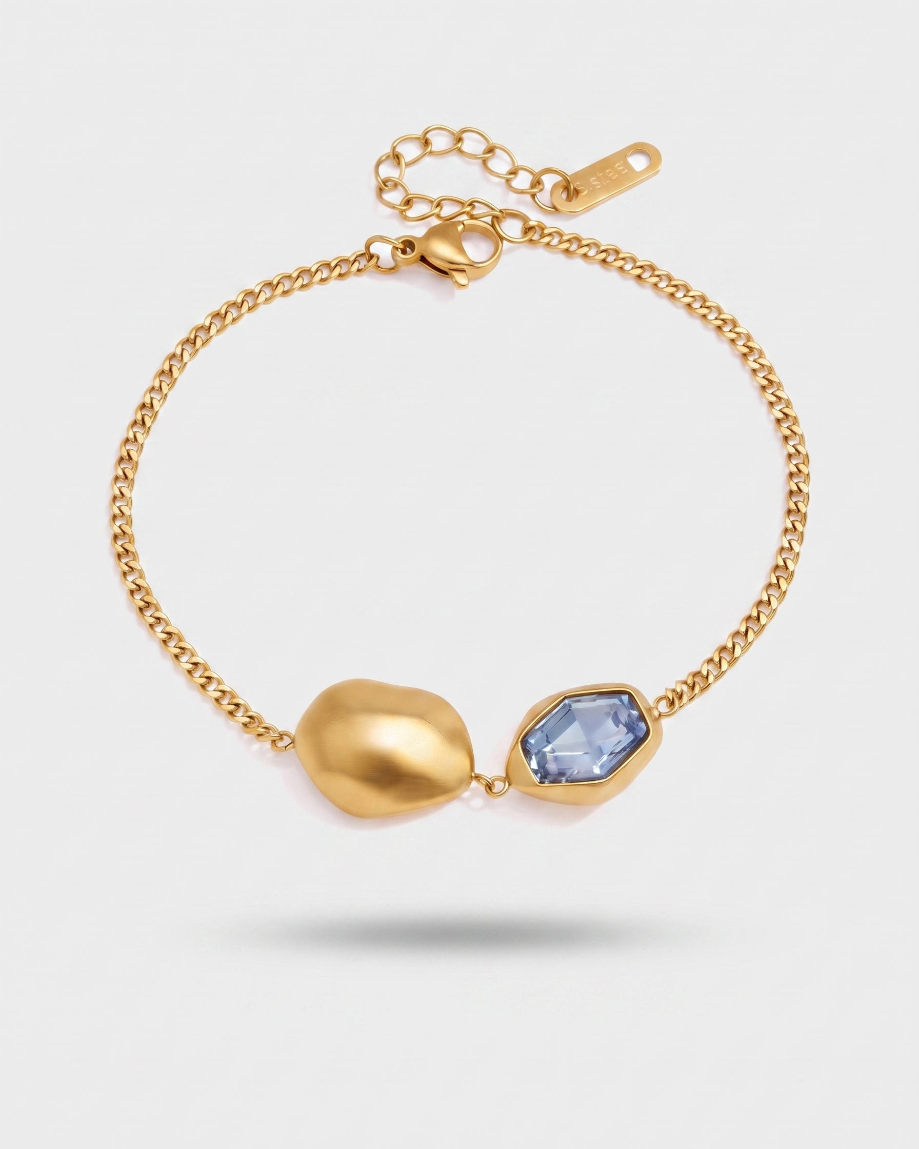 A gold chain bracelet featuring a large, organic-shaped gold bead and a faceted light blue gemstone in a bezel setting, displayed on a light gray background.