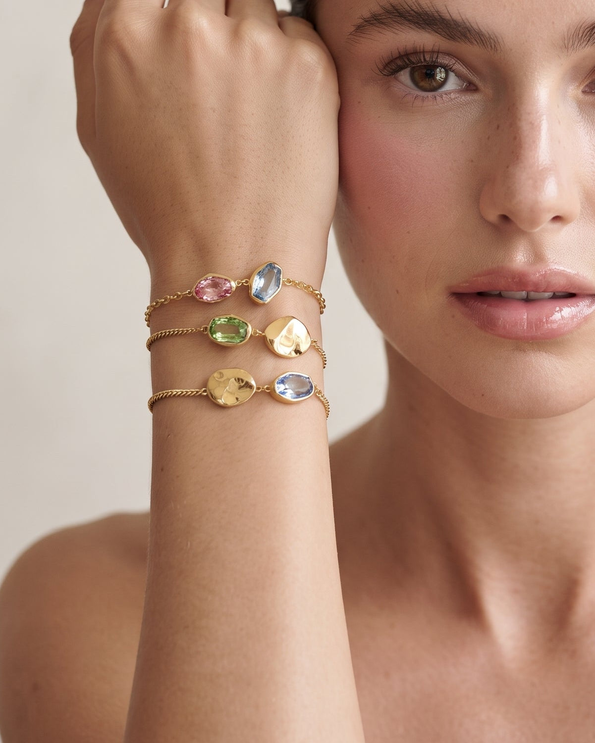 A close-up portrait of a woman wearing four delicate gold bracelets on her wrist, each featuring a different colored stone or charm, against a soft beige background.