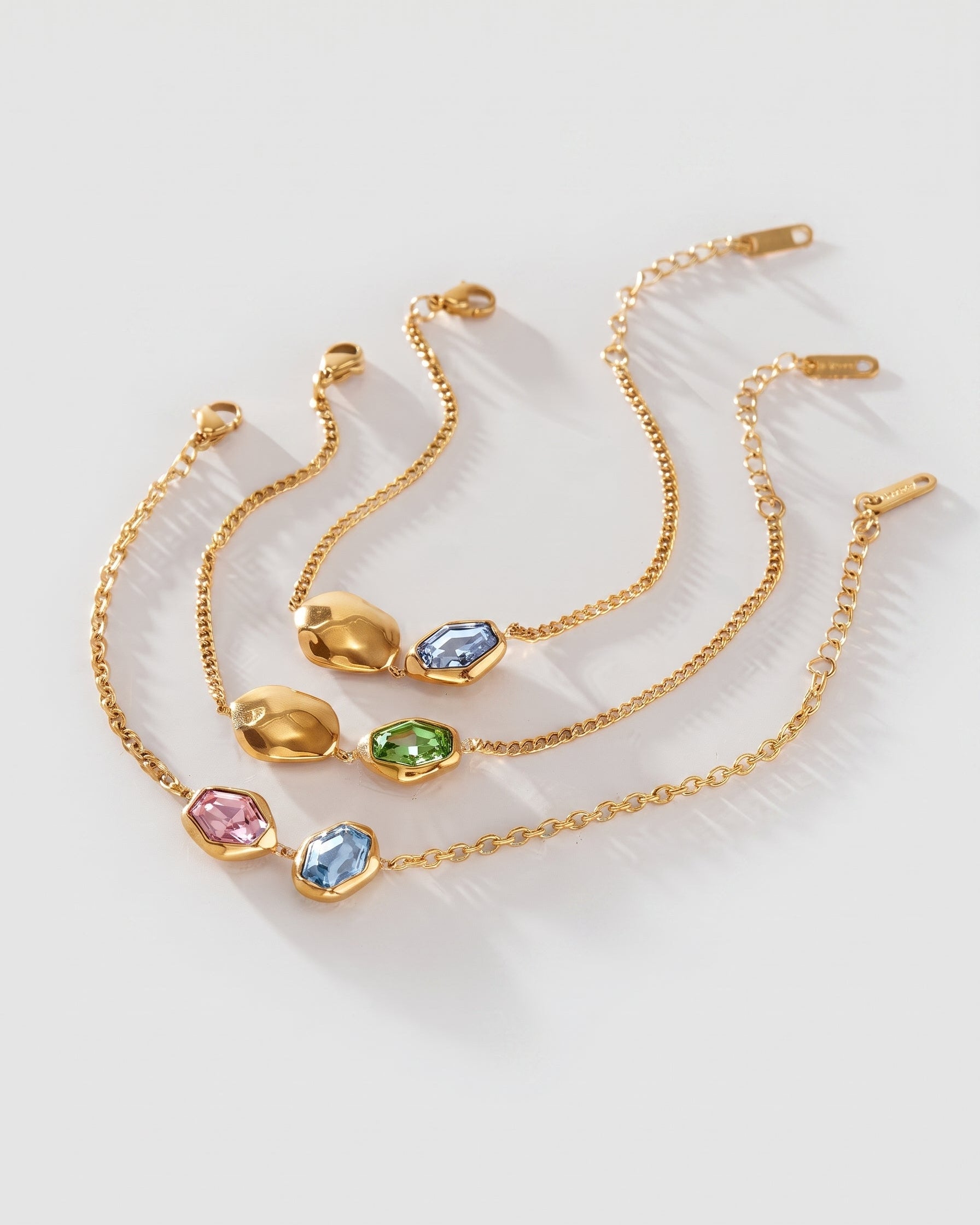 Three gold chain bracelets with irregular oval gemstone charms in pink, blue, and green, arranged on a light gray background.