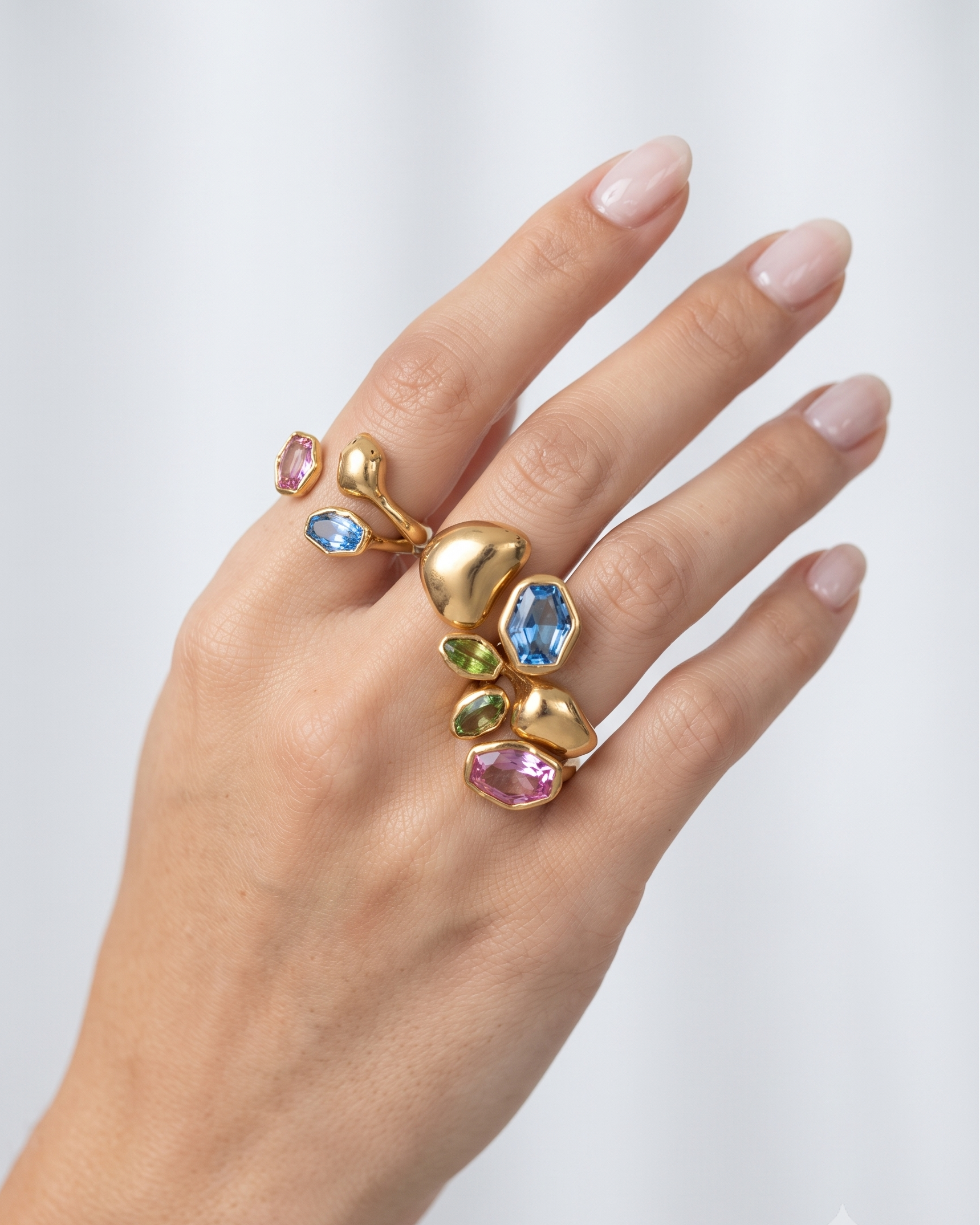 A close-up of a hand wearing a sculptural gold ring set with multiple colorful gemstones, including pink, blue, and green stones, against a soft light gray background.