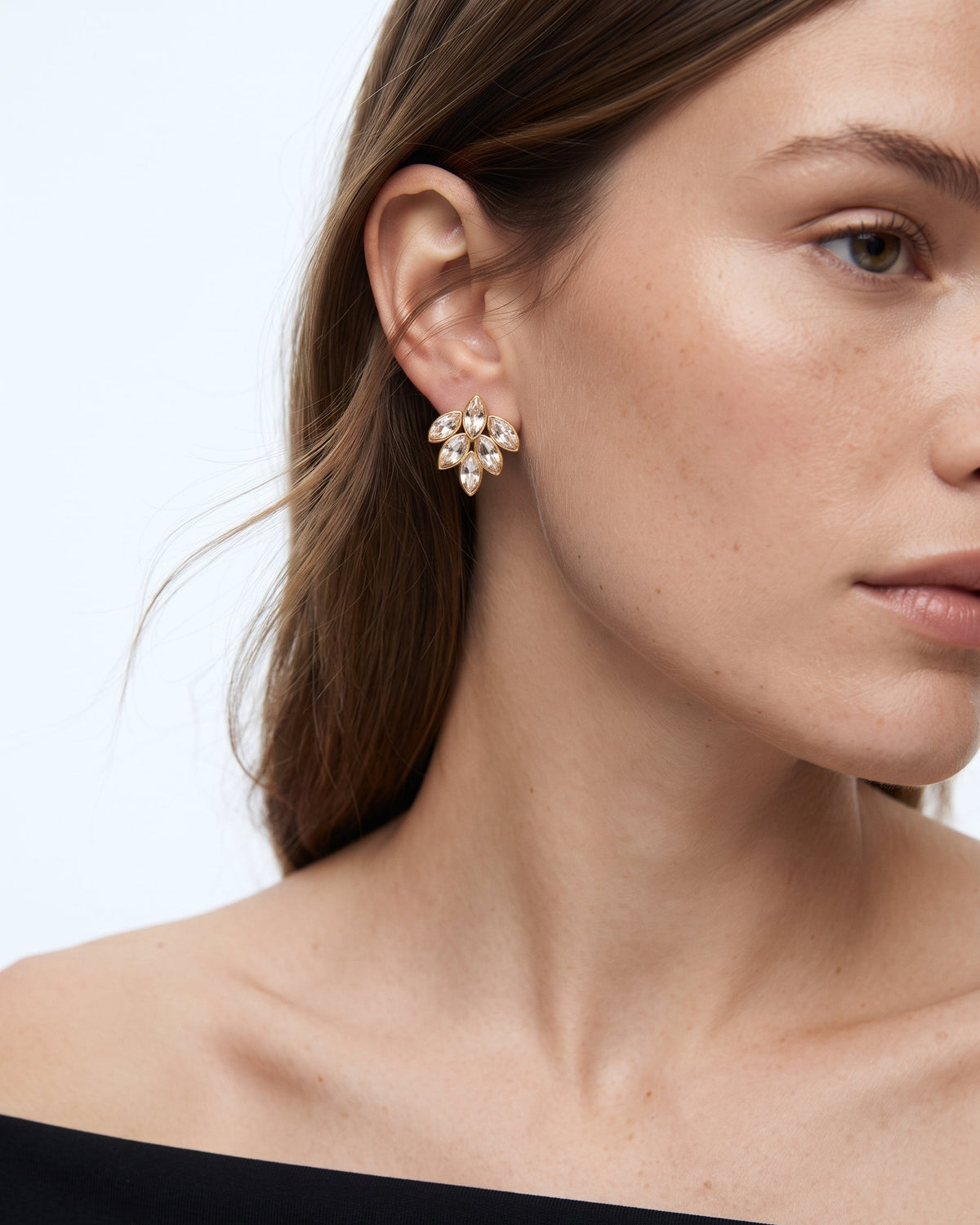 A close-up of a woman's ear and side profile wearing a small gold-toned floral cluster stud earring with clear marquise-cut stones against a light gray background.