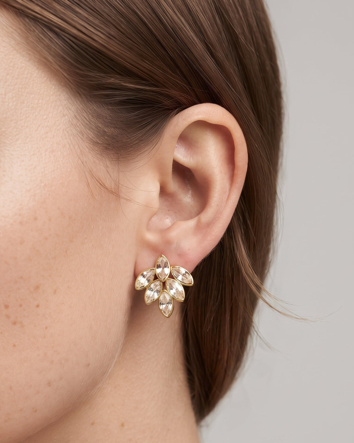 A close-up of a model's ear wearing a small gold-toned cluster stud earring with clear teardrop-shaped crystals, set against a plain light gray background.