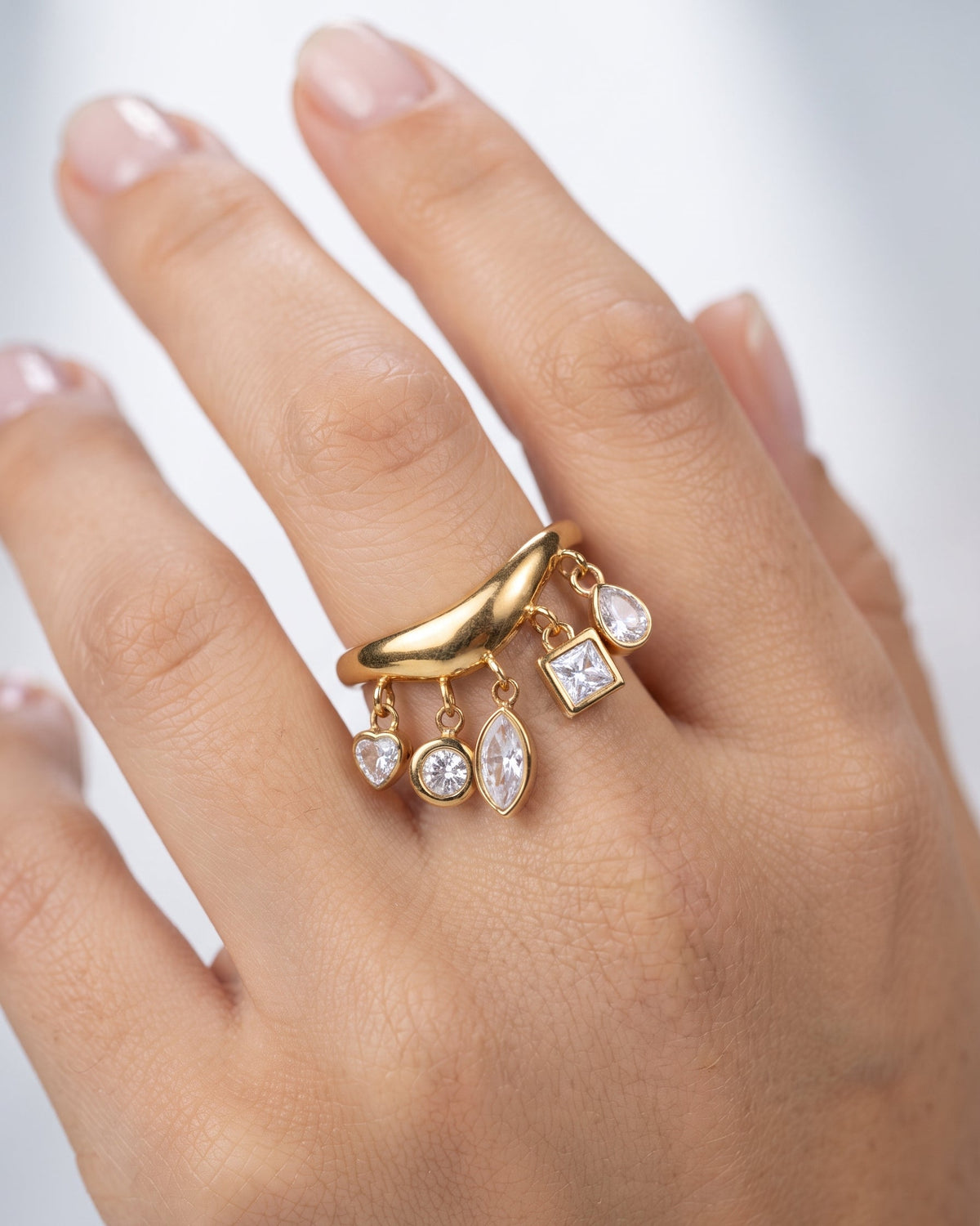 A gold ring with dangling clear gemstones, shown worn on a person's hand against a soft light gray background.