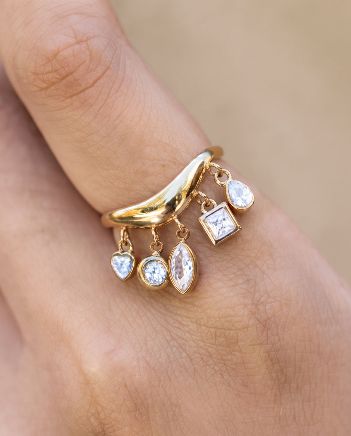 A gold crescent moon ring with dangling clear gemstones in heart, oval, marquise, and square settings, shown worn on a person's finger against a soft beige background.