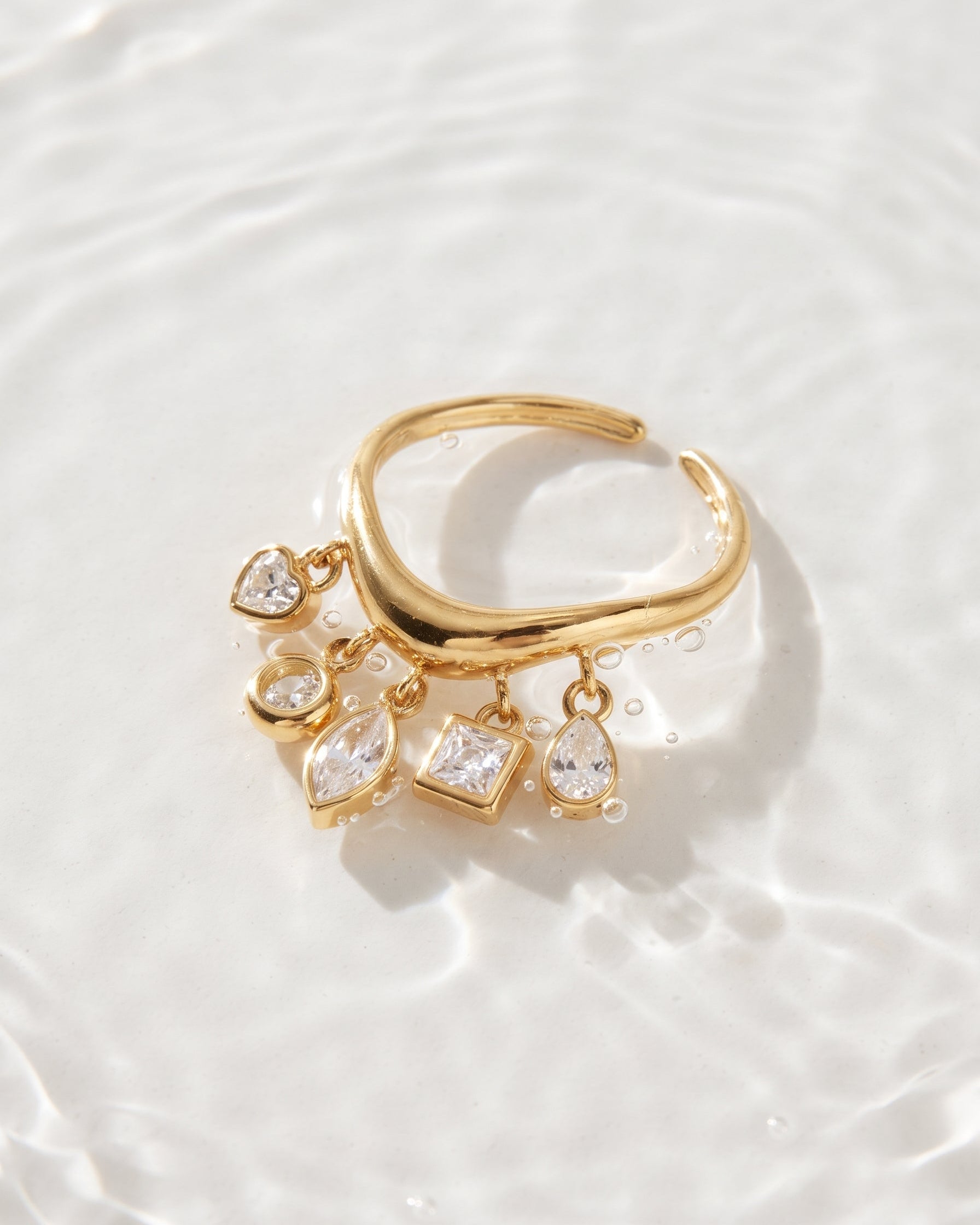 A gold-toned open cuff ring with dangling clear gemstone charms in heart, oval, pear, and square shapes, photographed on a softly rippled white surface.