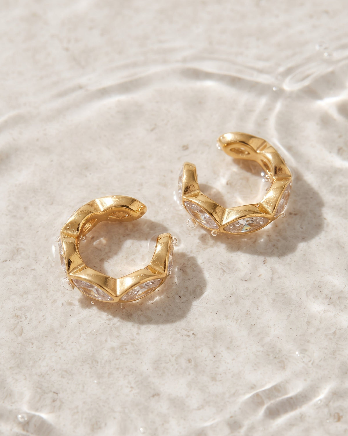 A pair of gold huggie hoop earrings with clear crystal accents, resting on a textured beige surface with water ripples in the background.
