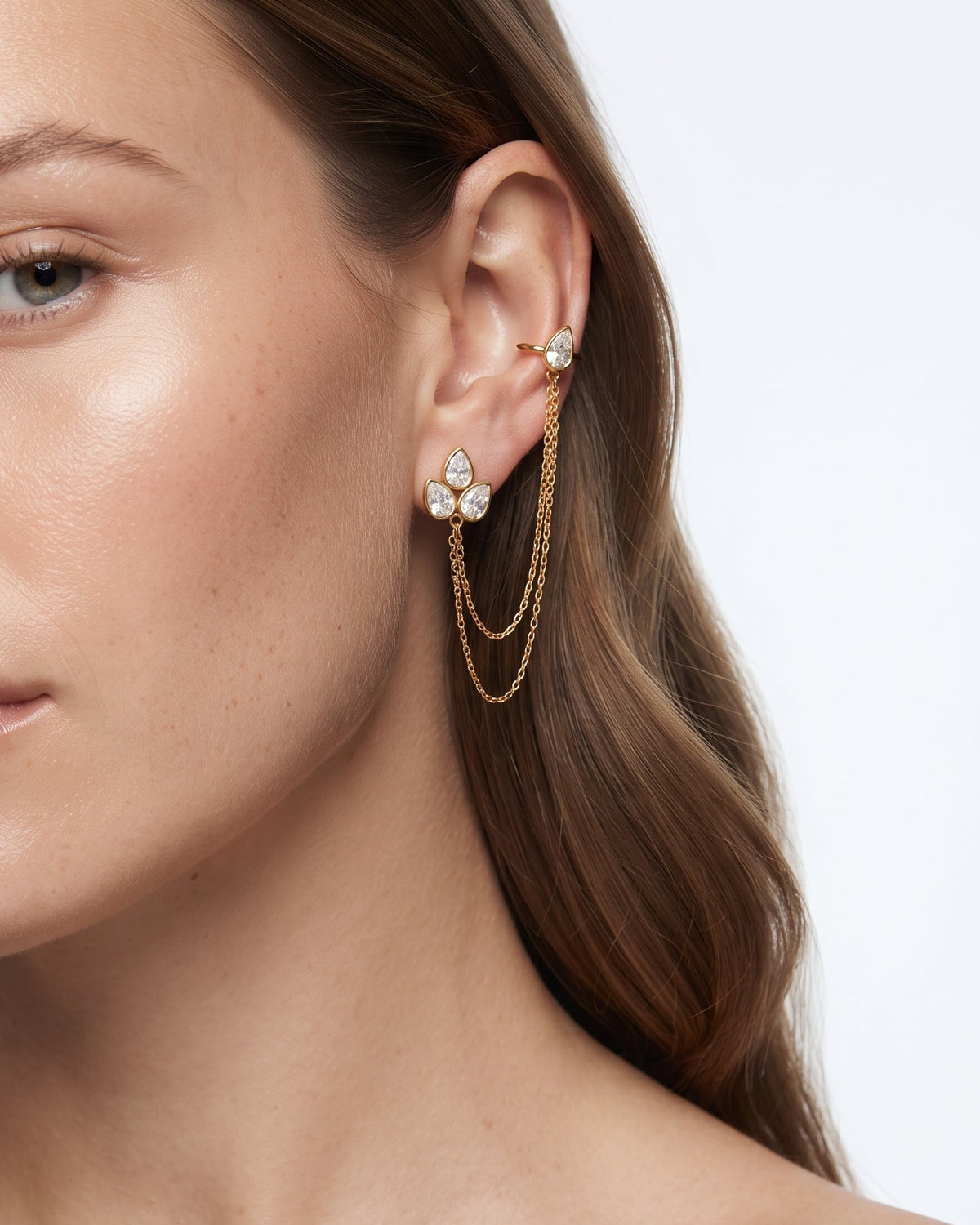A close-up of a woman's ear wearing a gold ear cuff with a floral stud earring and long dangling chain earrings, against a clean white background.
