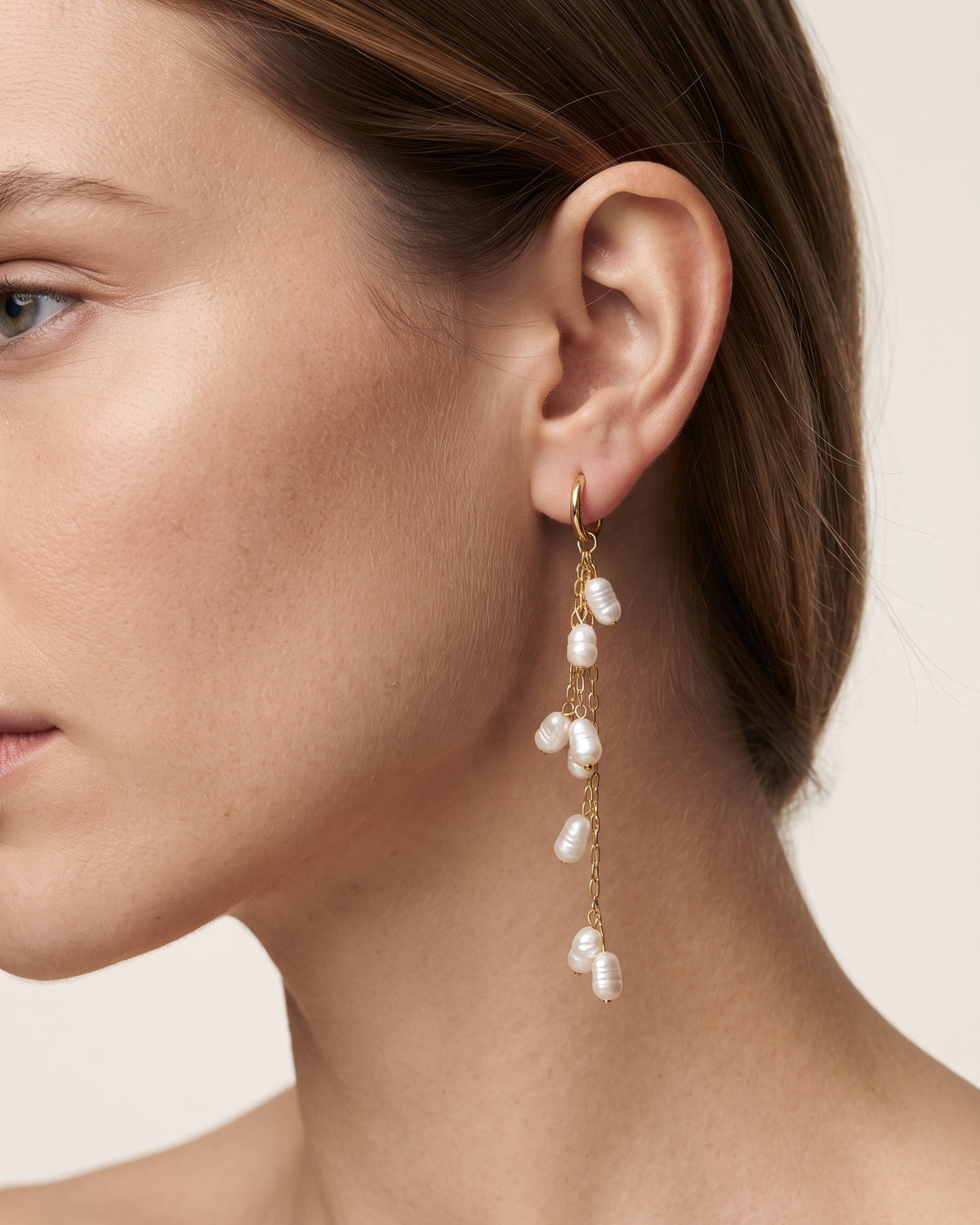 A close-up of a model's ear wearing a gold huggie hoop earring with a dangling gold chain adorned with irregular white baroque pearls.