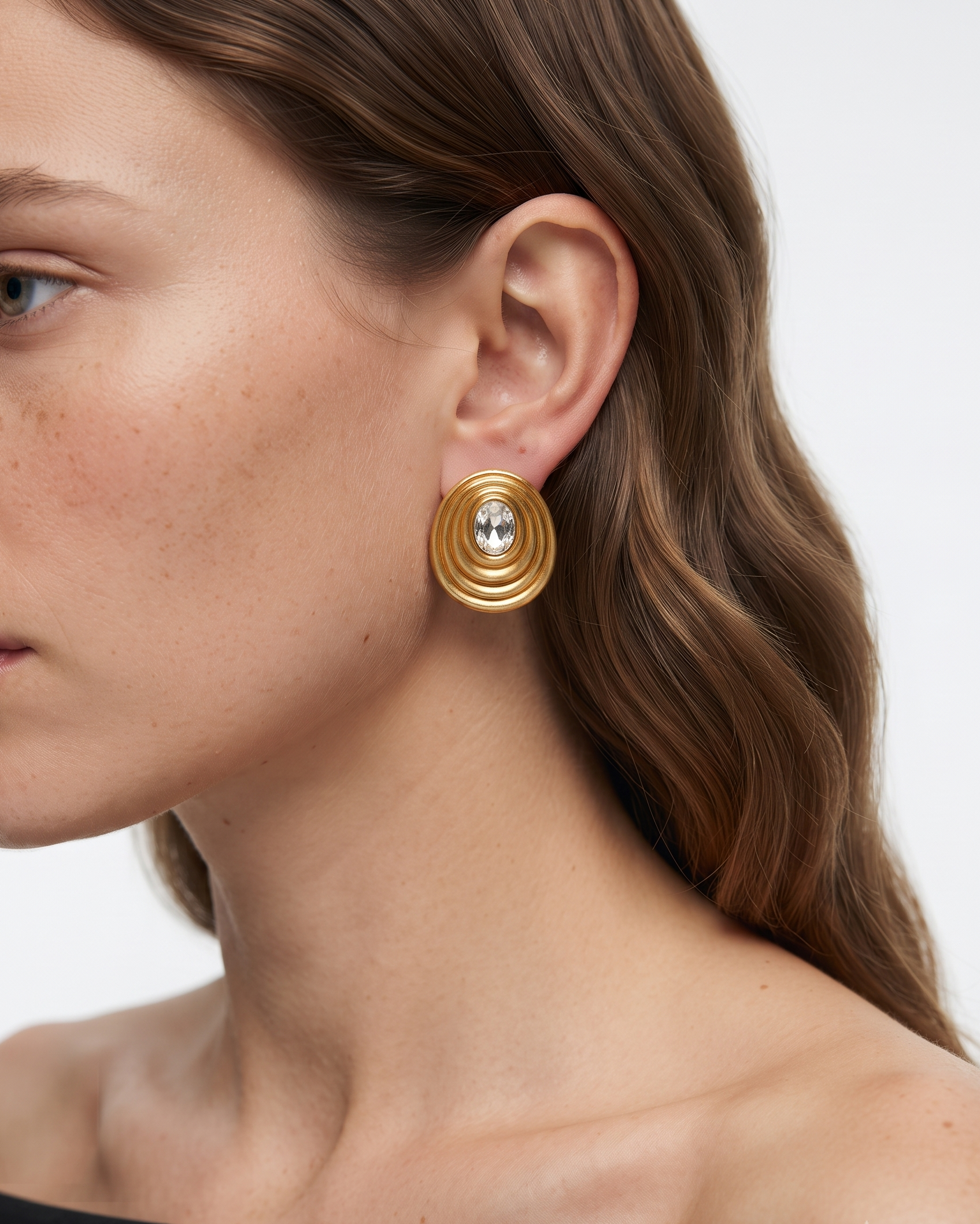 A close-up side profile of a woman wearing a gold-toned spiral earring with a clear central gemstone, against a clean light gray background.