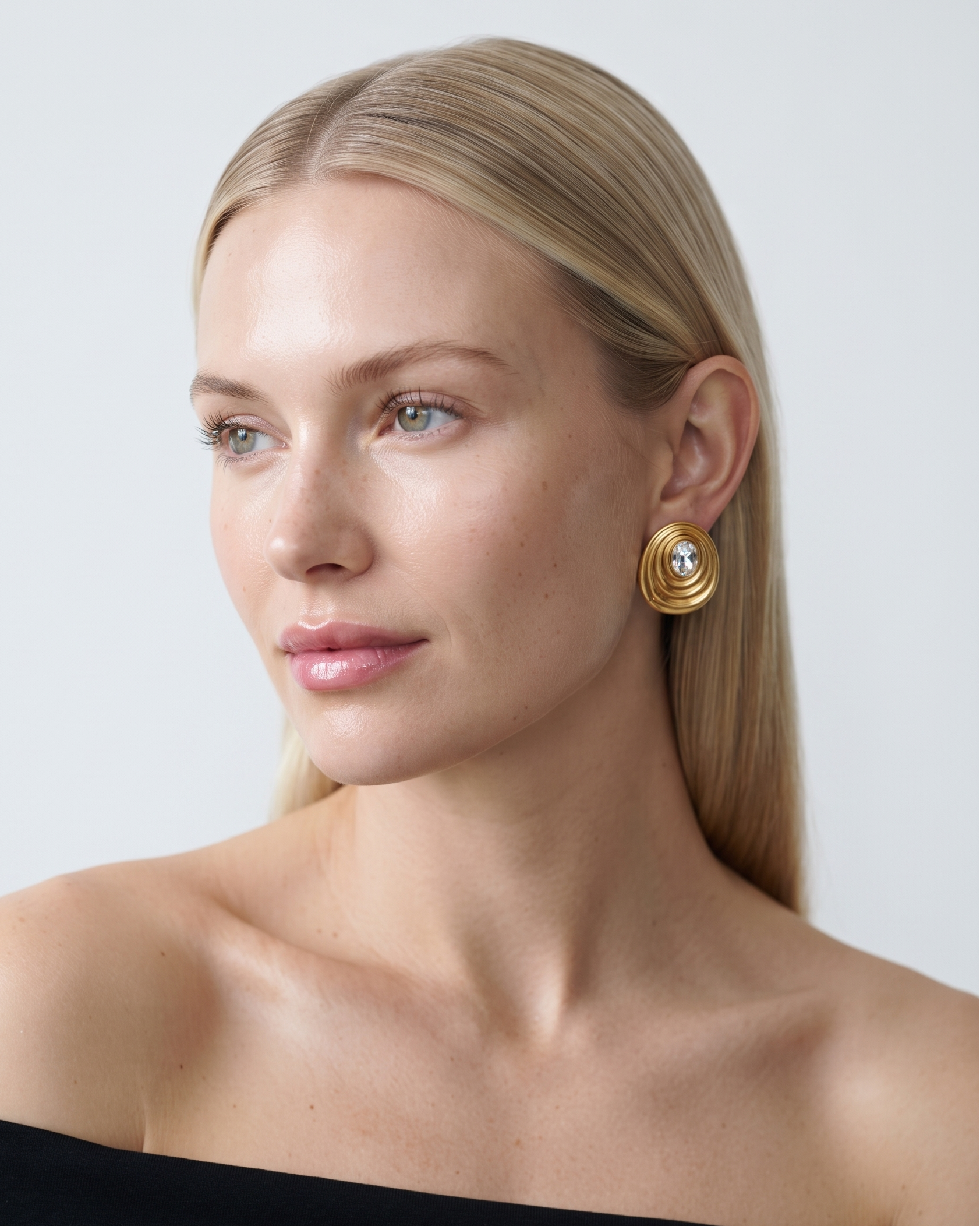 A close-up portrait of a blonde woman wearing a gold spiral drop earring with a central pearl, paired with a black off-the-shoulder top against a light gray background.