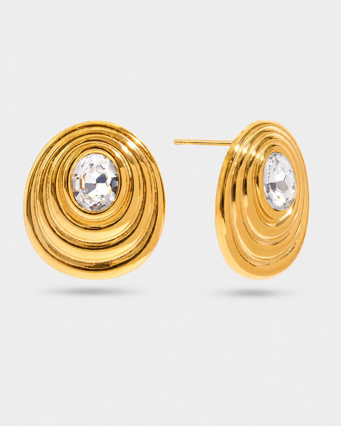 A pair of gold-toned disc earrings with concentric ridges and a clear faceted crystal in the center, shown on a light gray background.