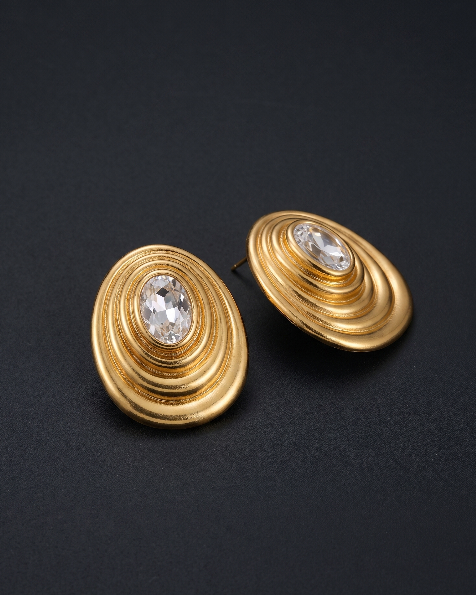 A pair of gold oval stud earrings with a clear faceted gemstone at the center, displayed on a dark textured background.