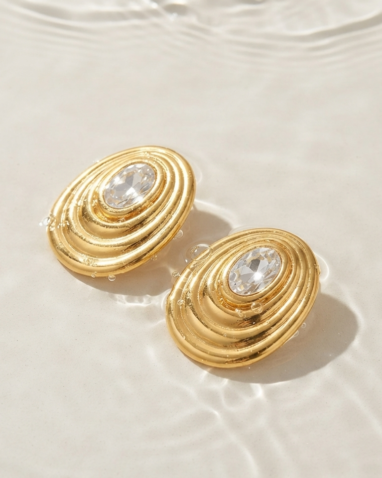 A pair of gold oval stud earrings with a clear faceted gemstone in the center, resting on a light beige surface with water ripples in the background.