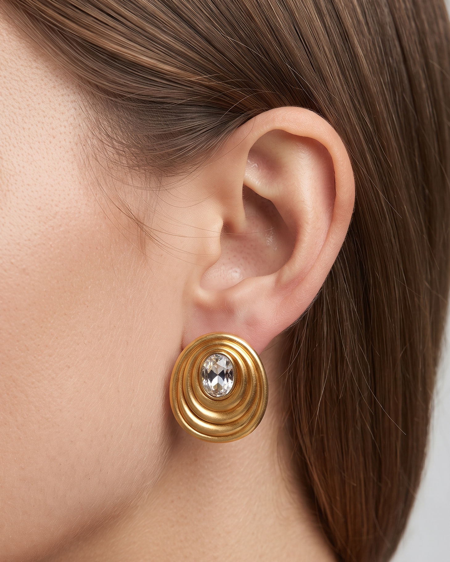 A close-up of a model's ear wearing a gold spiral stud earring with a clear central gemstone, set against a neutral gray background.
