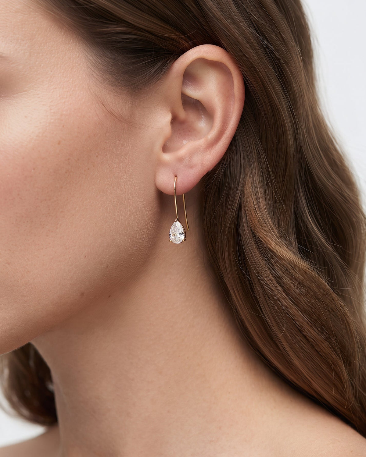 A close-up side profile of a woman wearing a delicate gold hoop earring with a small clear teardrop gemstone dangle, set against a clean white background.
