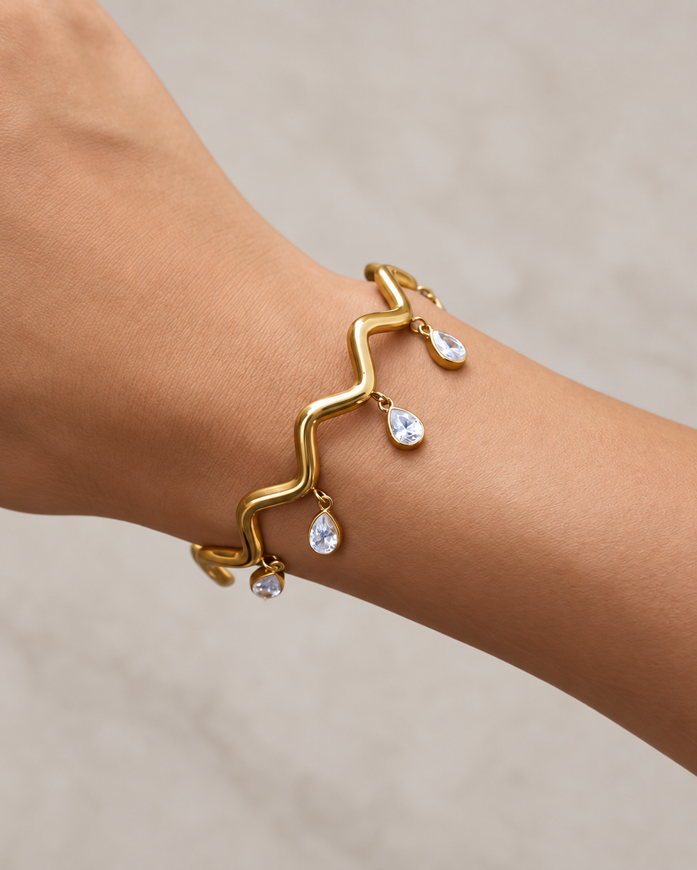 A gold wavy bracelet with three dangling clear teardrop gemstones, shown worn on a person's wrist against a light beige background.