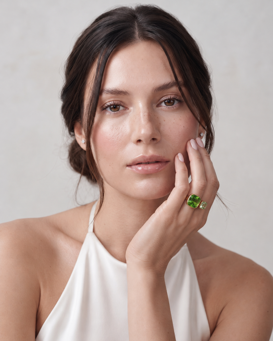 A woman with dark hair pulled back wears a white halter top and a green gemstone ring on her finger, posing against a light gray background.