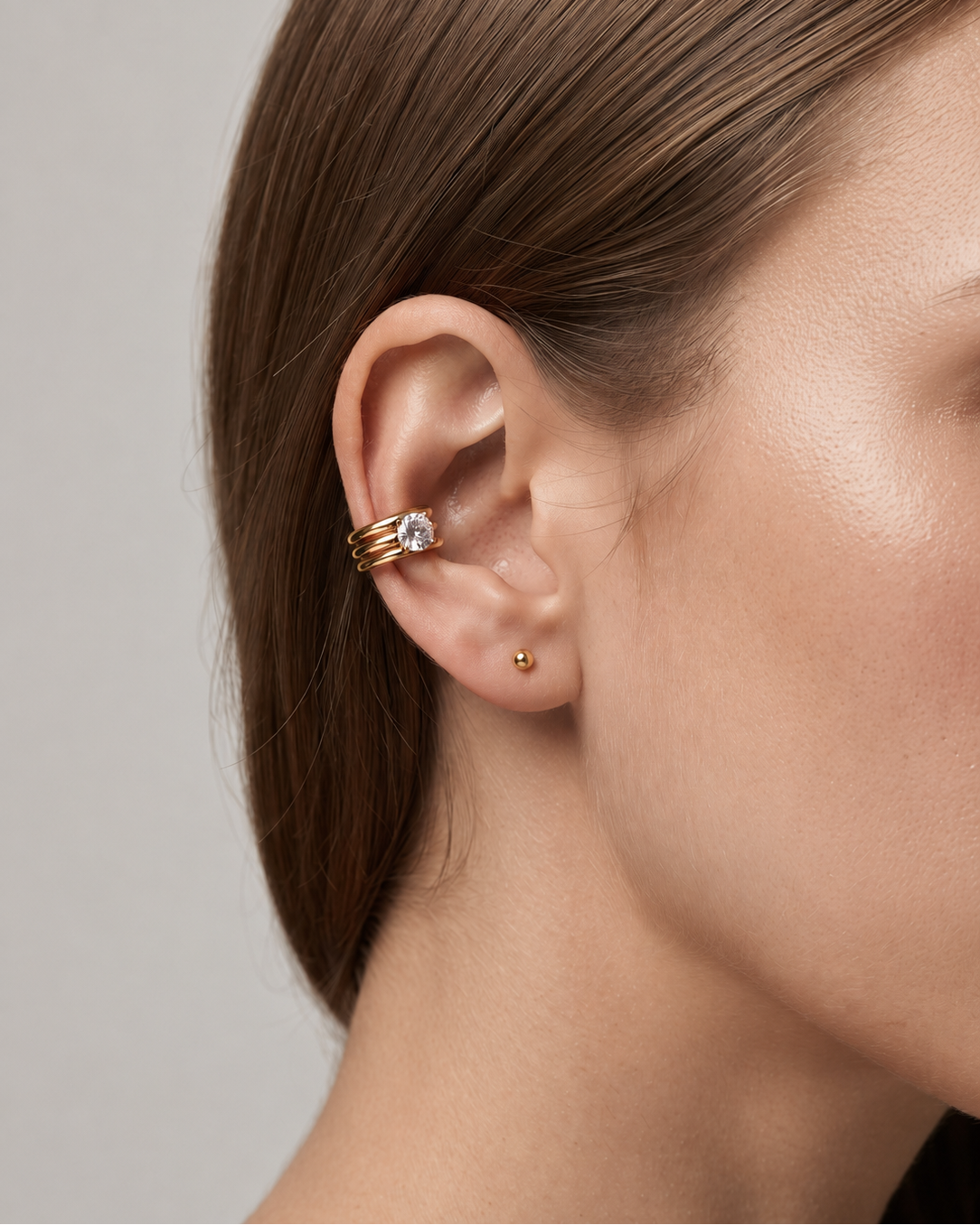 A close-up of a model's ear wearing a gold ear cuff with a clear gemstone, along with a small gold stud earring, against a neutral gray background.