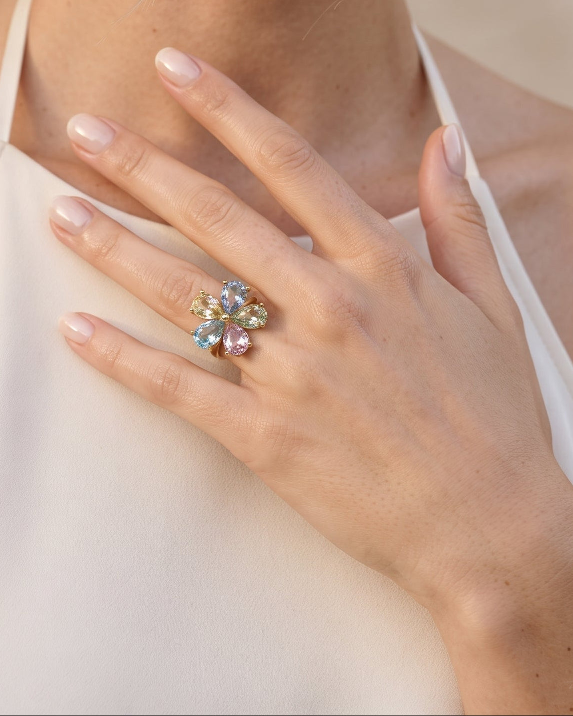 A close-up lifestyle shot of a woman wearing a large, colorful floral ring on her finger, wearing a cream halter top against a soft beige background.