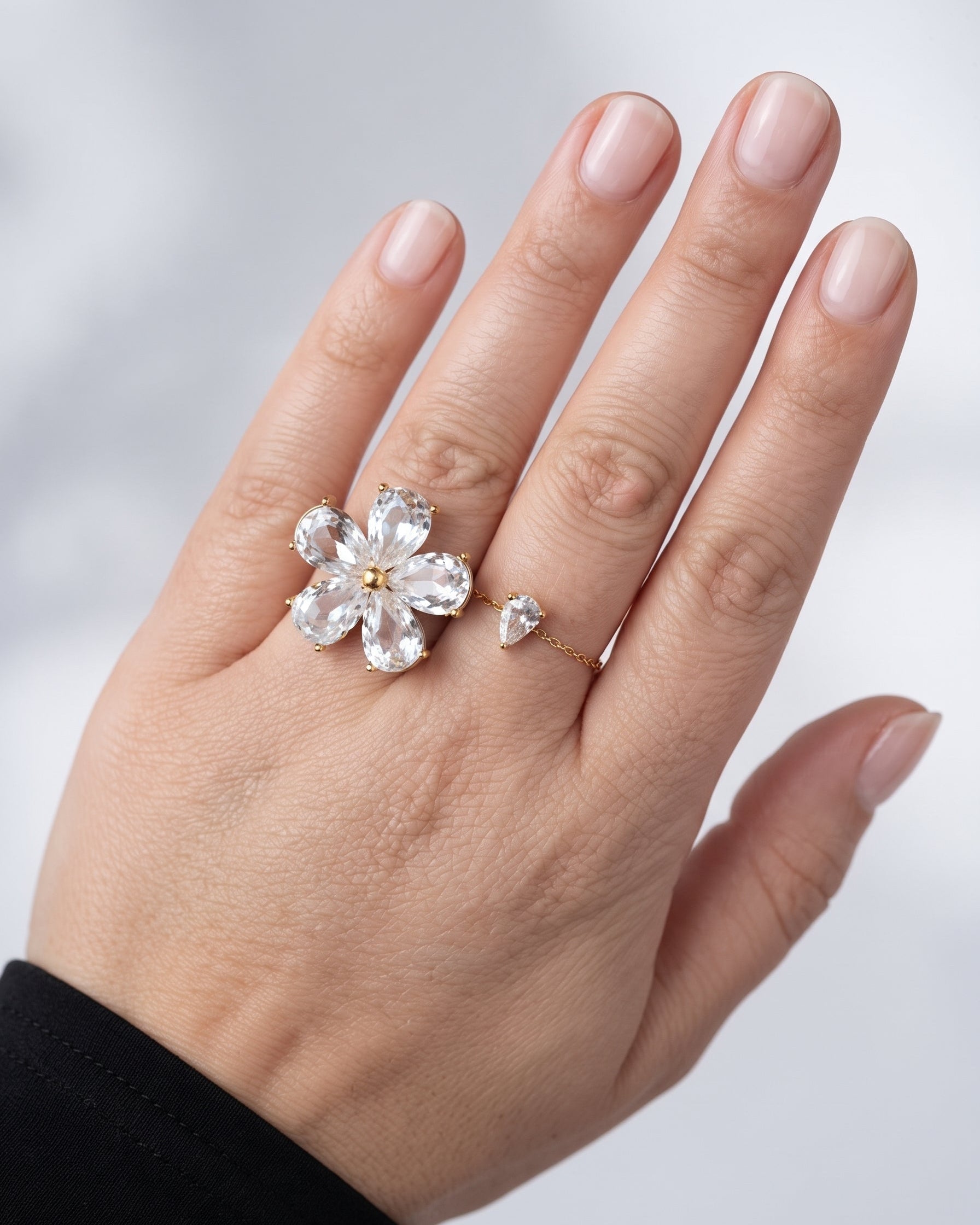 A close-up of a hand wearing a gold ring set with a clear crystal flower and a small matching crystal heart, against a soft light gray background.