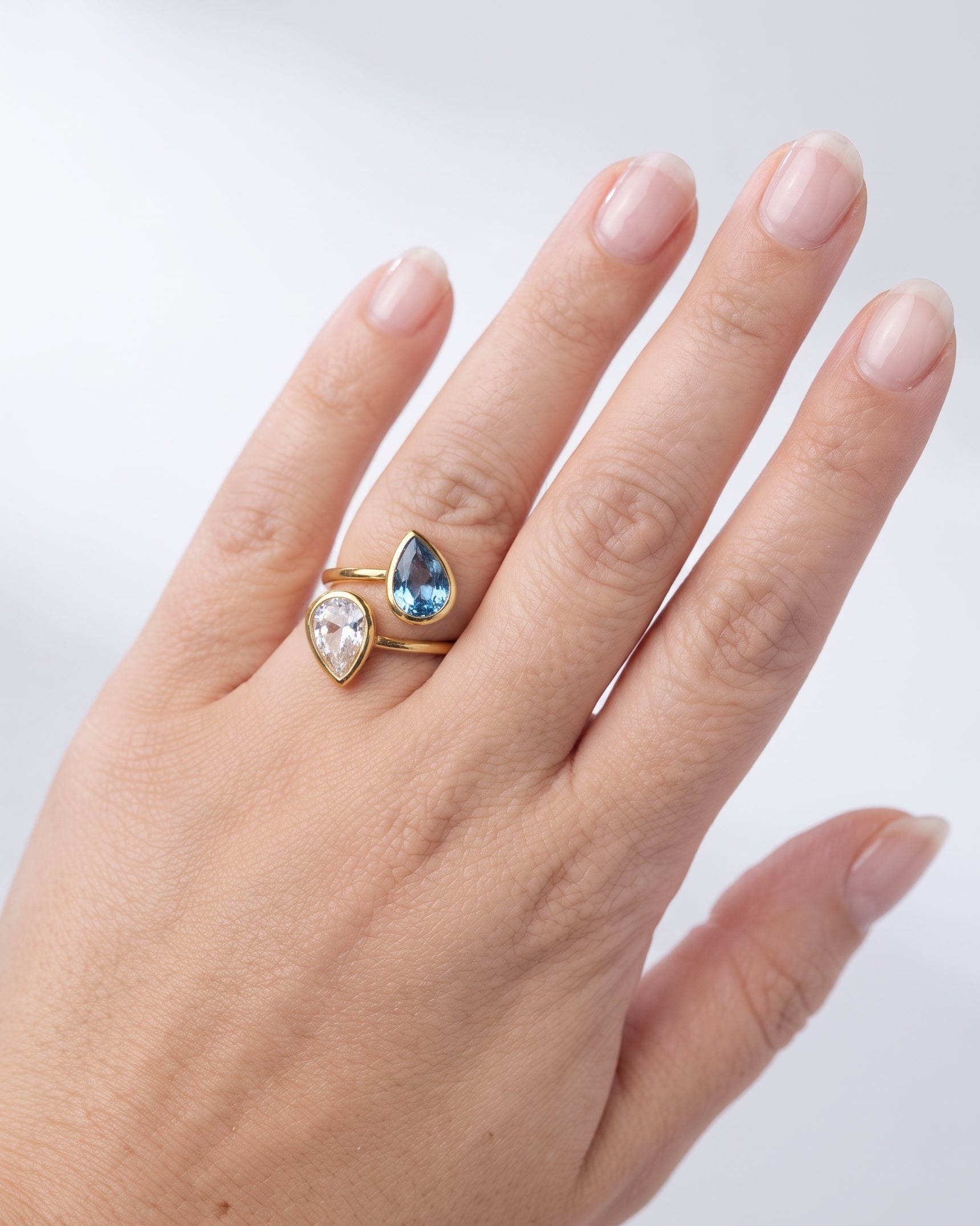 A gold ring with a pear-cut blue gemstone and a pear-cut clear gemstone, shown worn on a hand against a light gray background.