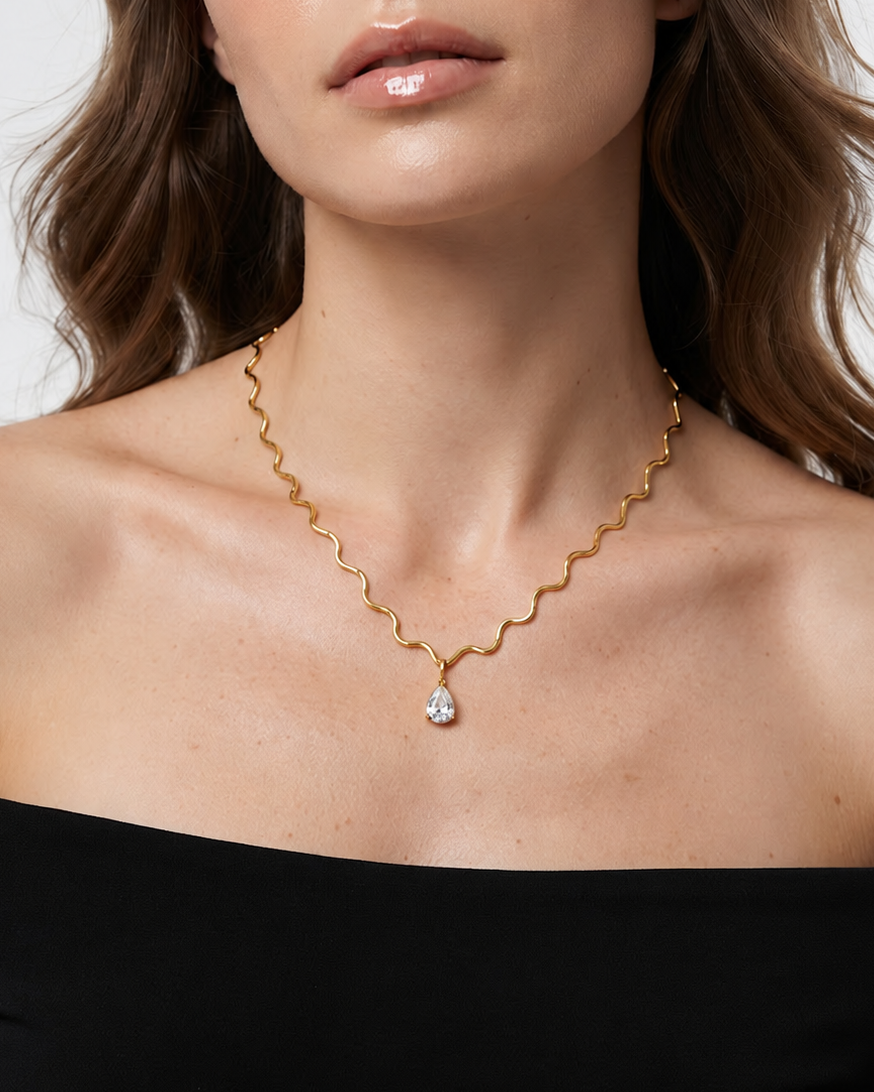 A woman wearing a delicate gold wavy chain necklace with a teardrop-shaped clear gemstone pendant, styled with a black off-the-shoulder top against a light gray background.