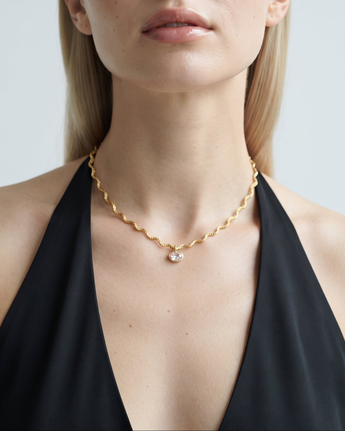 A blonde woman wearing a delicate gold chain necklace with a small white pearl pendant, styled with a black halter top against a light gray background.