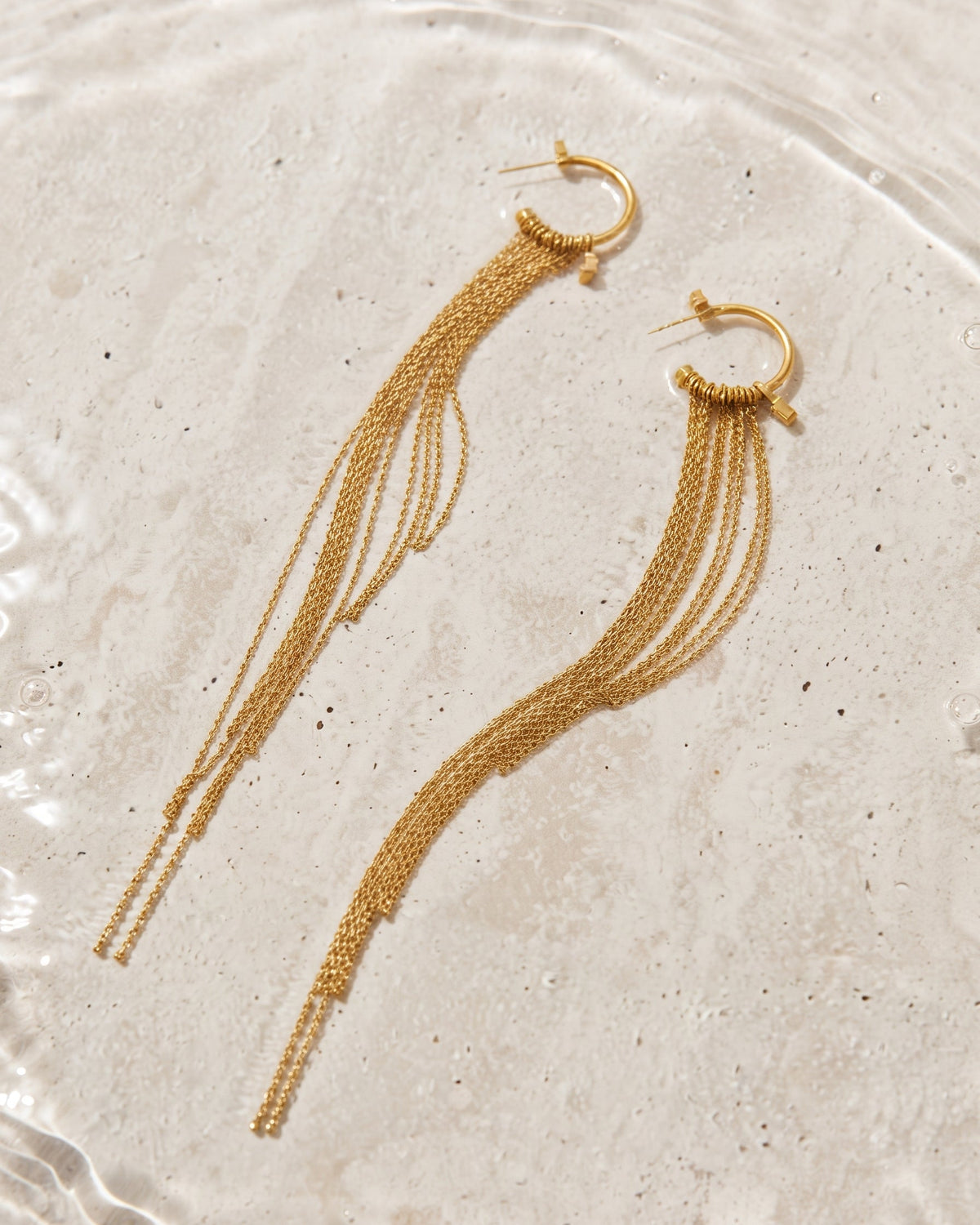 A pair of gold-toned dangle earrings with thick and thin chain strands, displayed on a textured light beige stone surface.