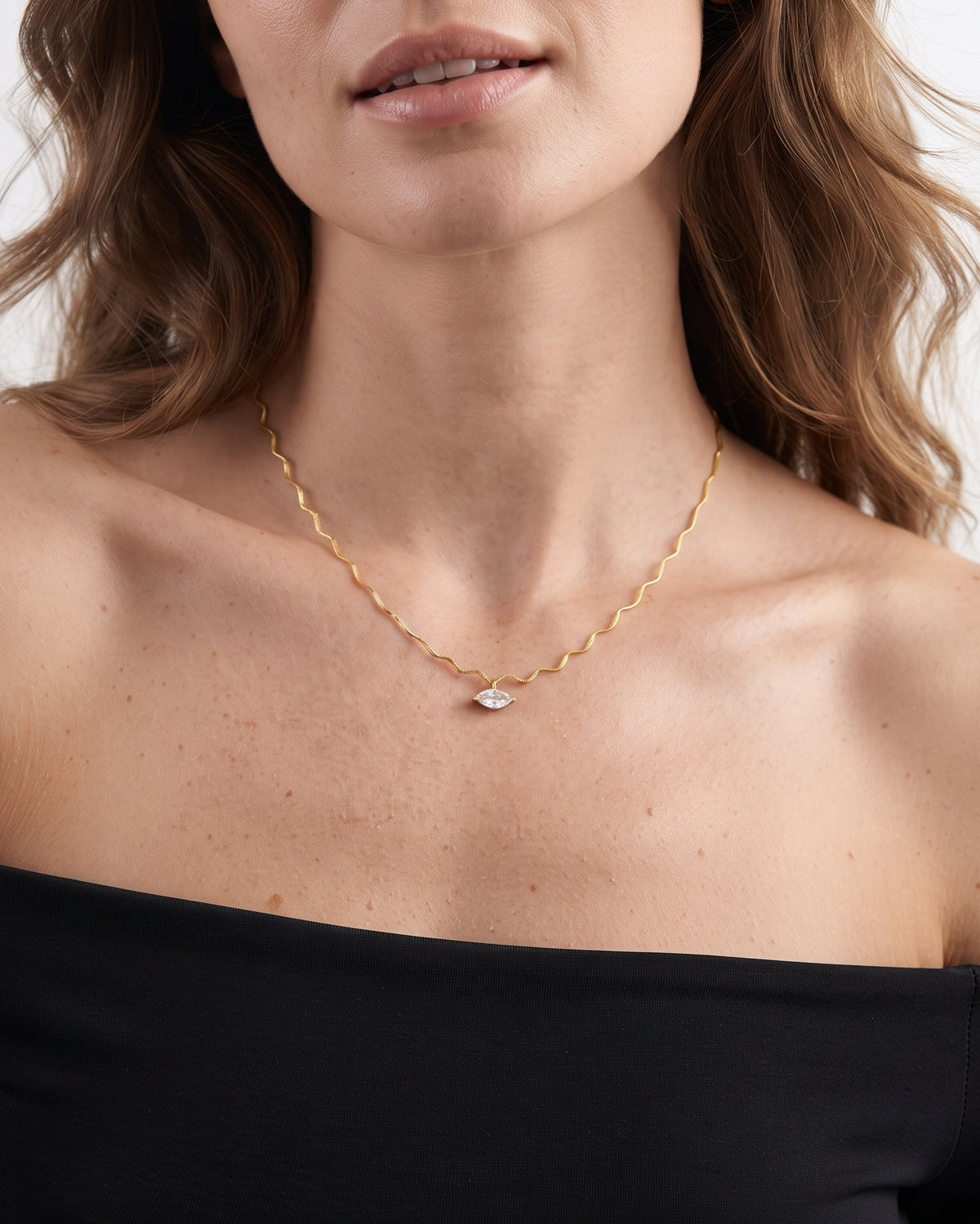 A close-up of a woman wearing a delicate gold chain necklace with a small white teardrop-shaped pendant, styled with a black off-the-shoulder top against a light gray background.