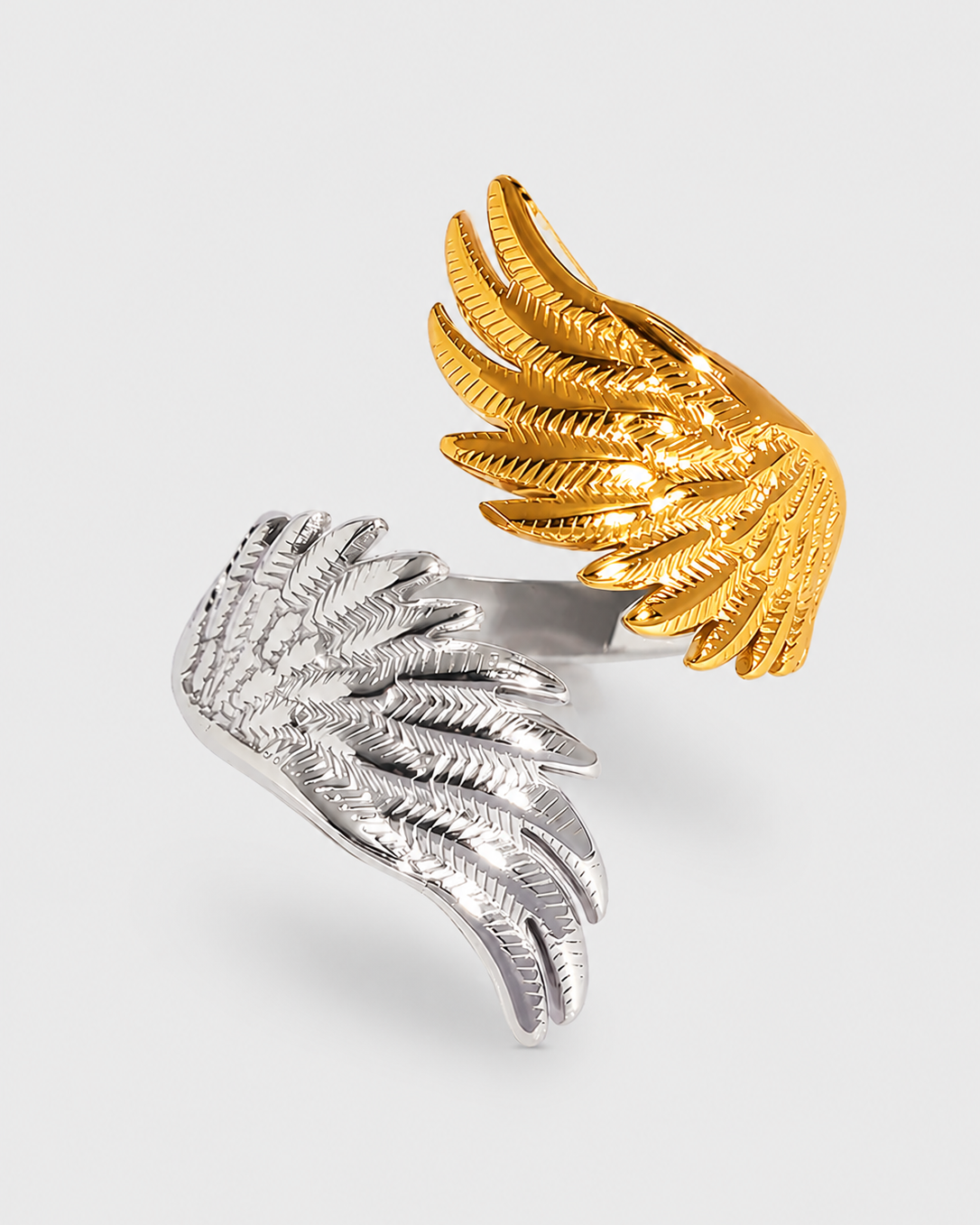 A sculptural wing-shaped ring featuring two textured wings, one in polished gold and one in polished silver, set against a light gray background.