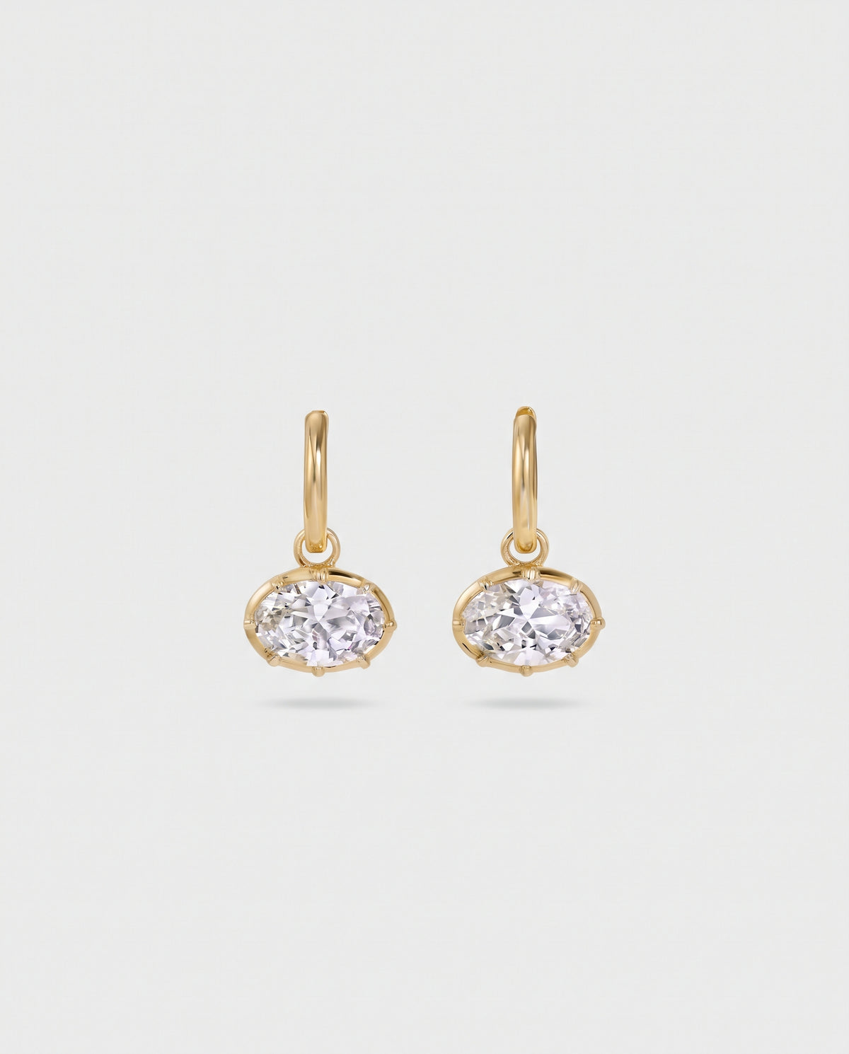 A pair of gold hoop earrings with oval bezel-set clear gemstones, displayed on a clean, light gray background.