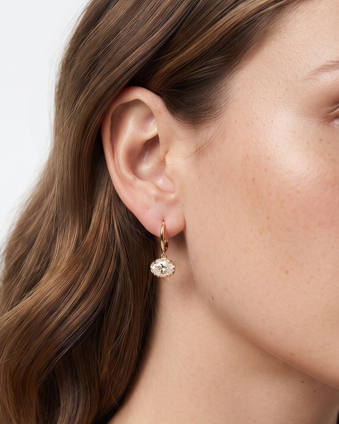 A close-up of a woman wearing a delicate gold dangle earring with a clear faceted gemstone, shown against a clean white background.