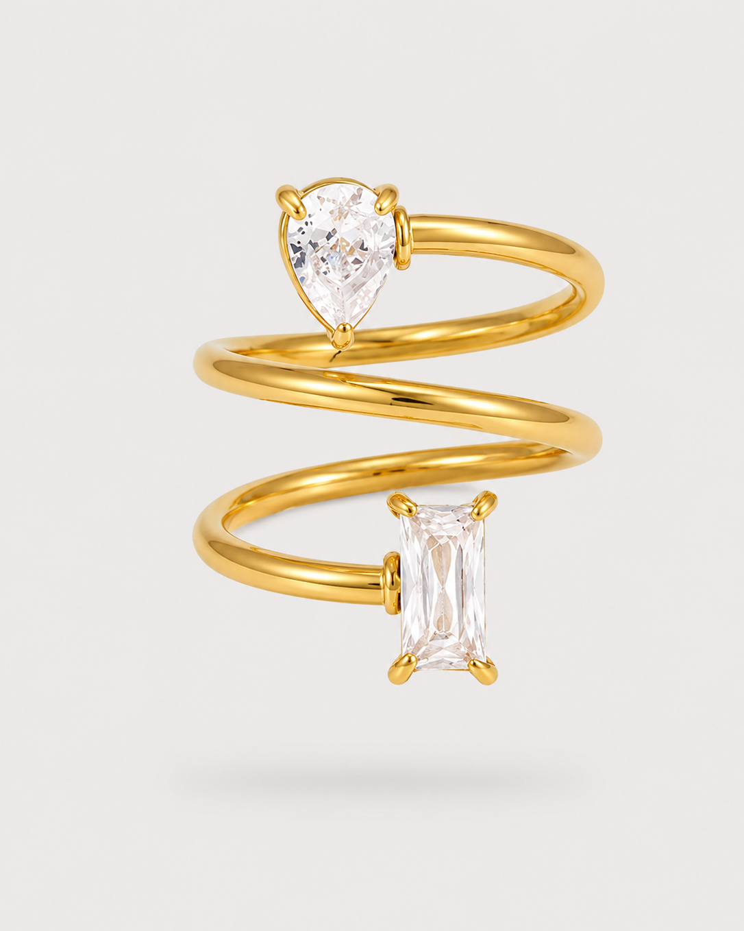 A gold spiral ring featuring a pear-cut clear gemstone on the upper spiral and an emerald-cut clear gemstone on the lower spiral, set against a light gray background.