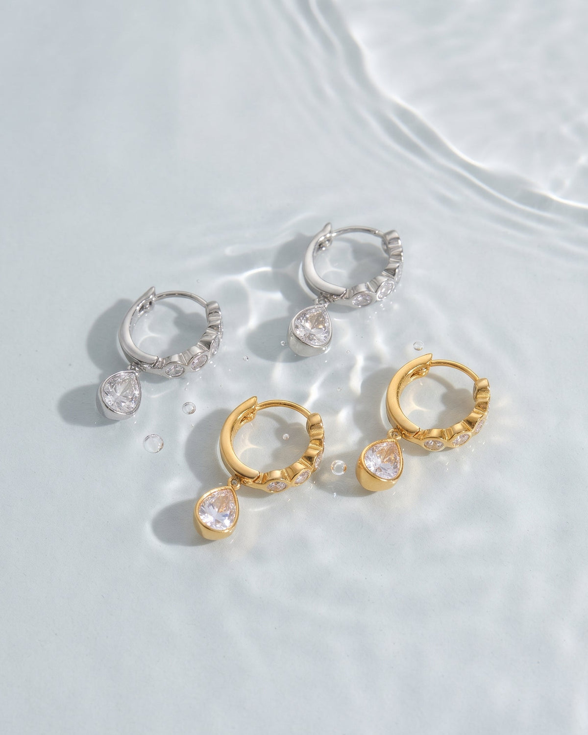 Three pairs of small hoop earrings with teardrop-shaped clear gemstones are arranged on a light, water-textured surface. Two pairs are gold-toned and one is silver-toned.