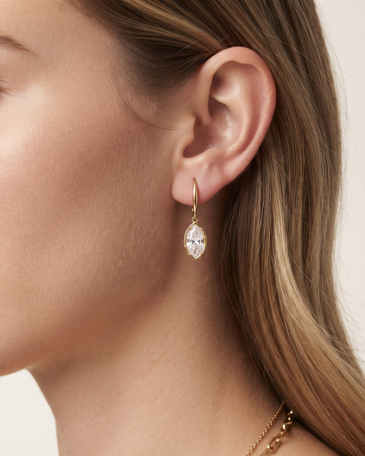 A close-up of a woman's ear wearing a gold drop earring with a marquise-cut clear gemstone, shown against her skin and light brown hair.