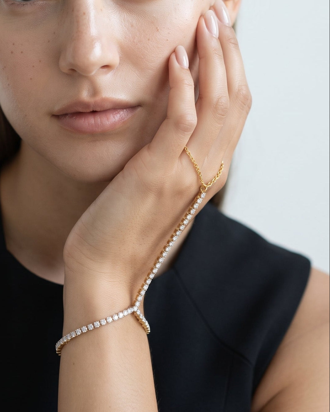 A close-up portrait of a woman with her hand resting against her cheek, wearing a delicate gold chain bracelet draped across her wrist and a matching gold ring, set against a clean light gray background.