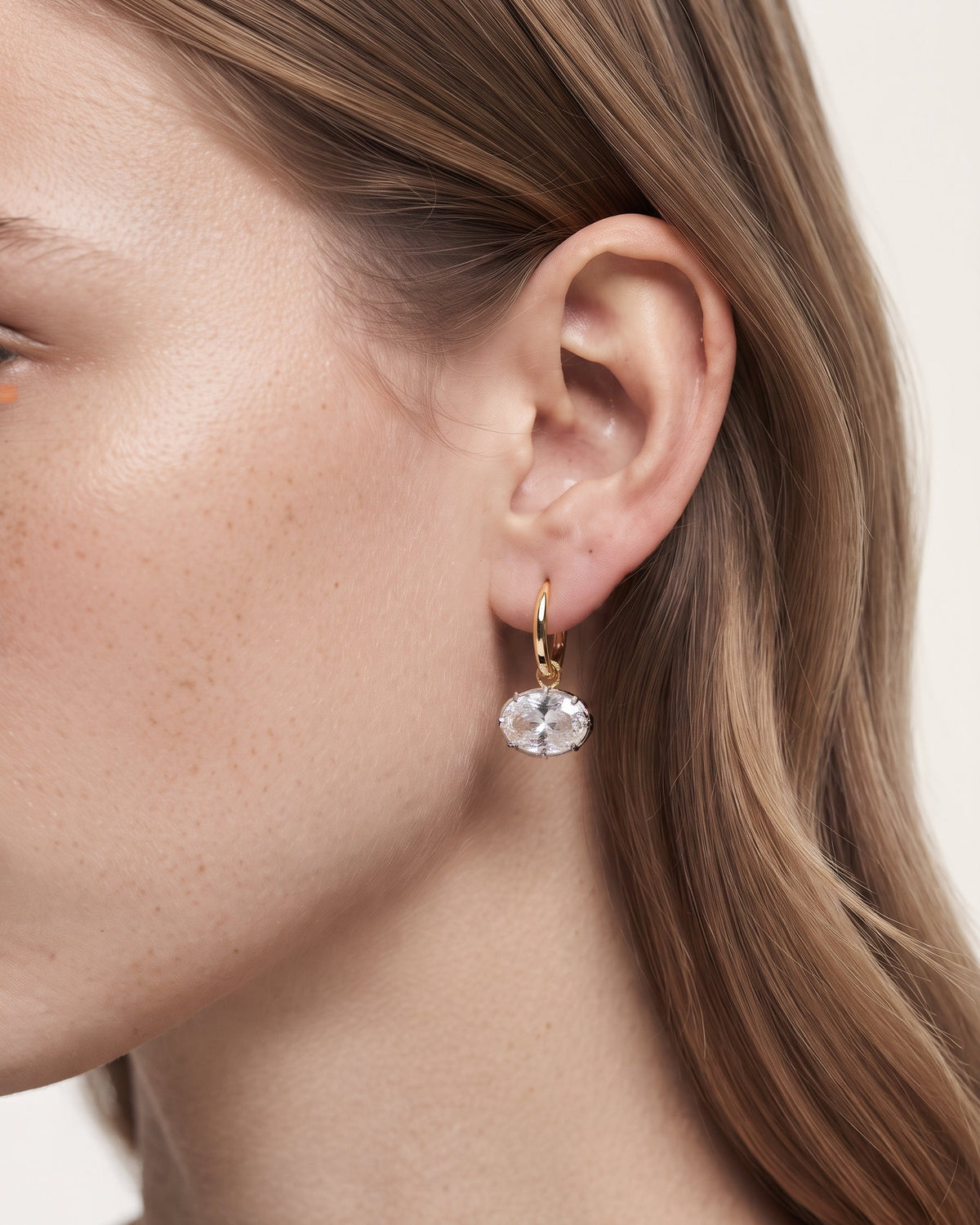 A close-up of a model's ear wearing a delicate gold huggie hoop earring with a clear faceted oval gemstone.
