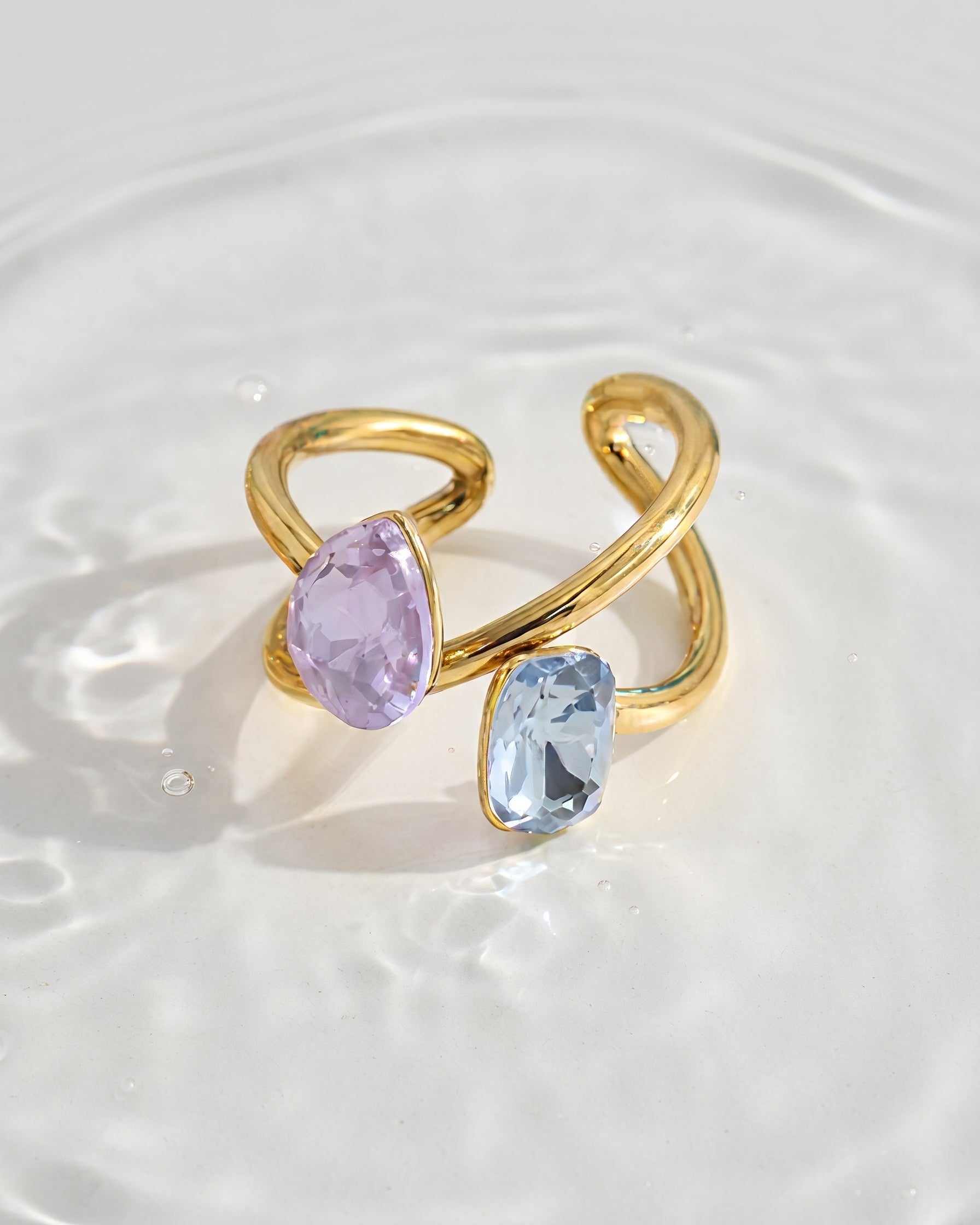 A gold-toned open wrap ring featuring two marquise-cut gemstones, one light purple and one light blue, resting on a surface of rippling water.