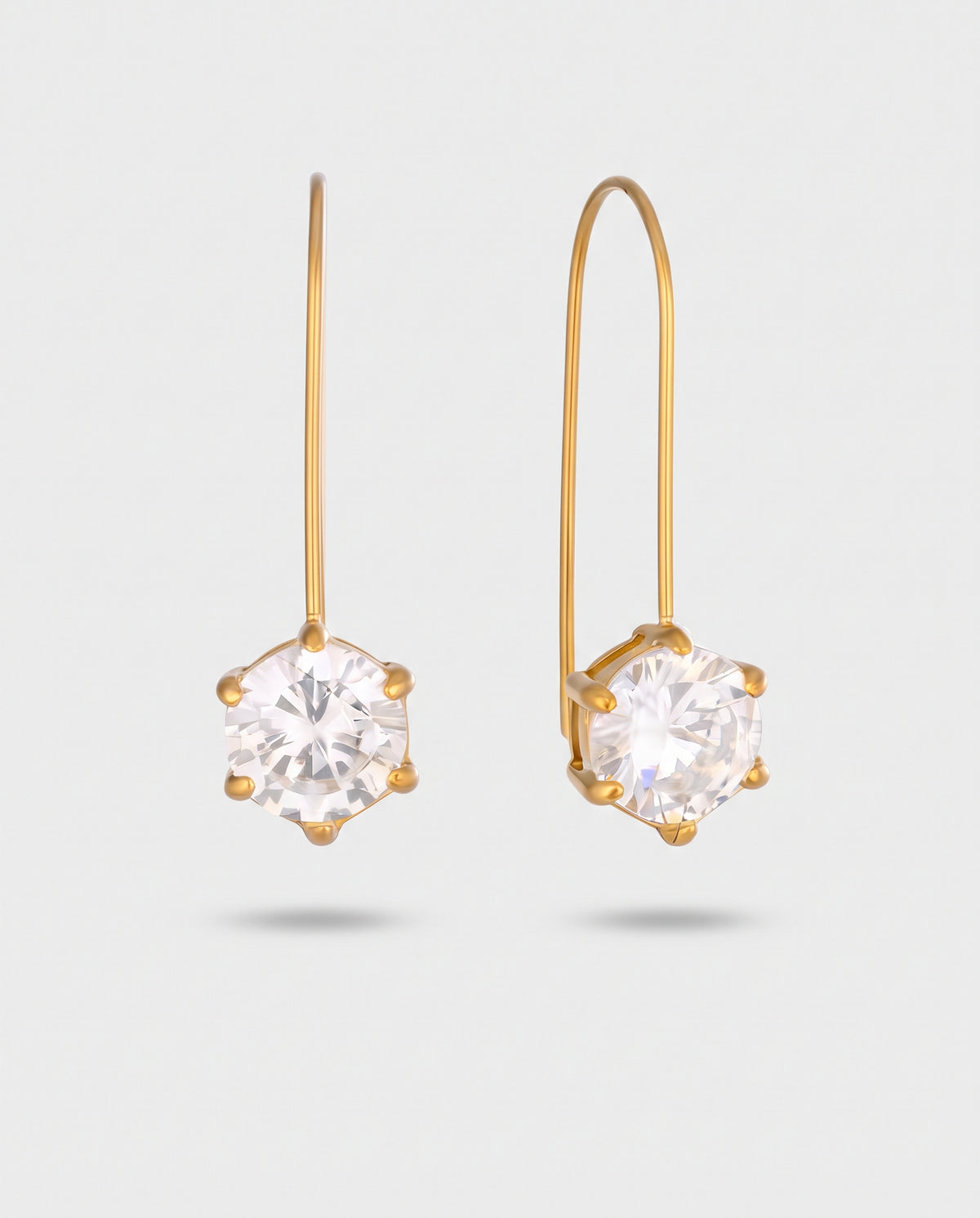 A pair of gold drop earrings with a single round clear gemstone in a prong setting, suspended from thin gold hooks against a light gray background.