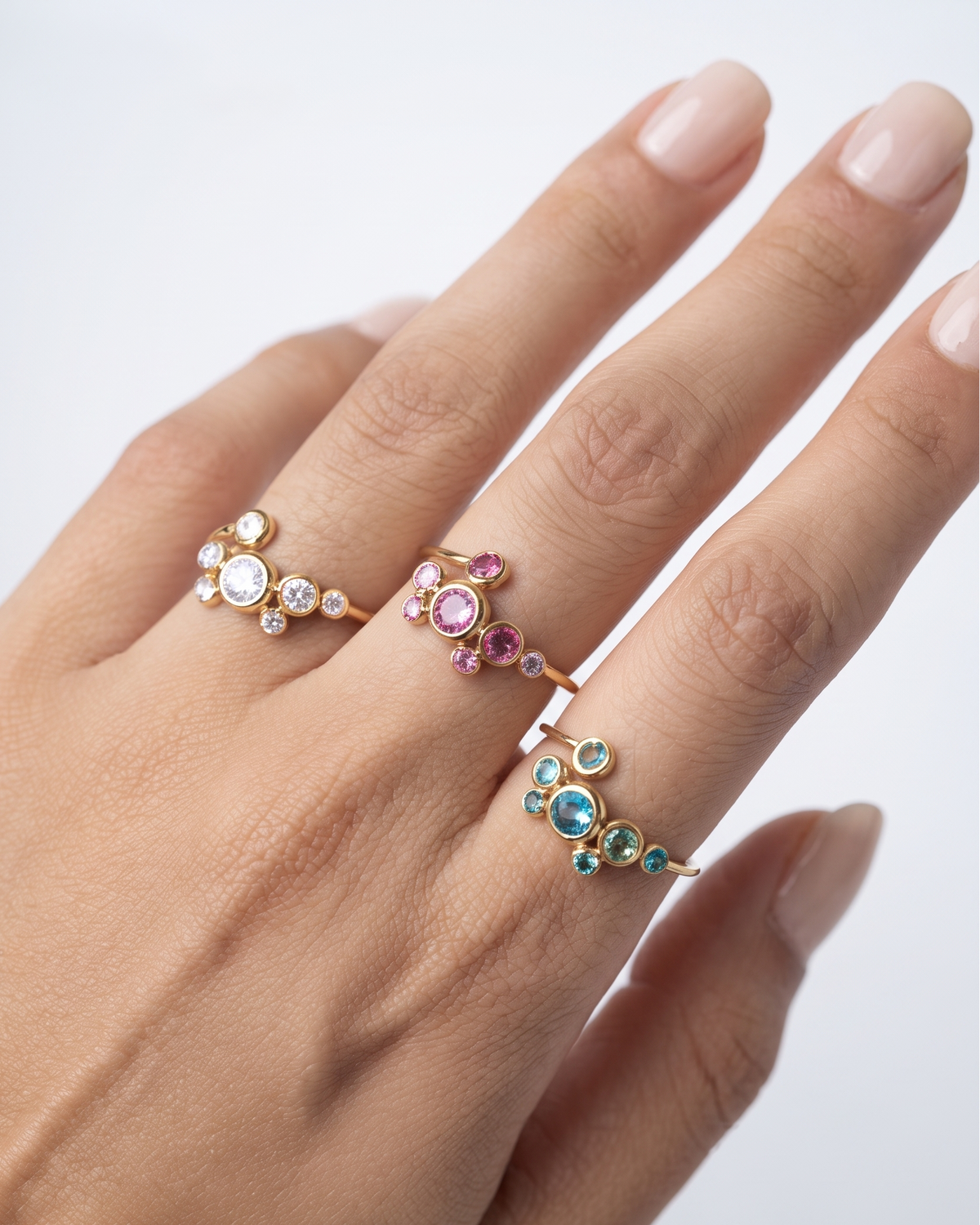A close-up of a hand wearing three delicate gold rings with small gemstones in clear, pink, blue, and green settings, against a clean white background.