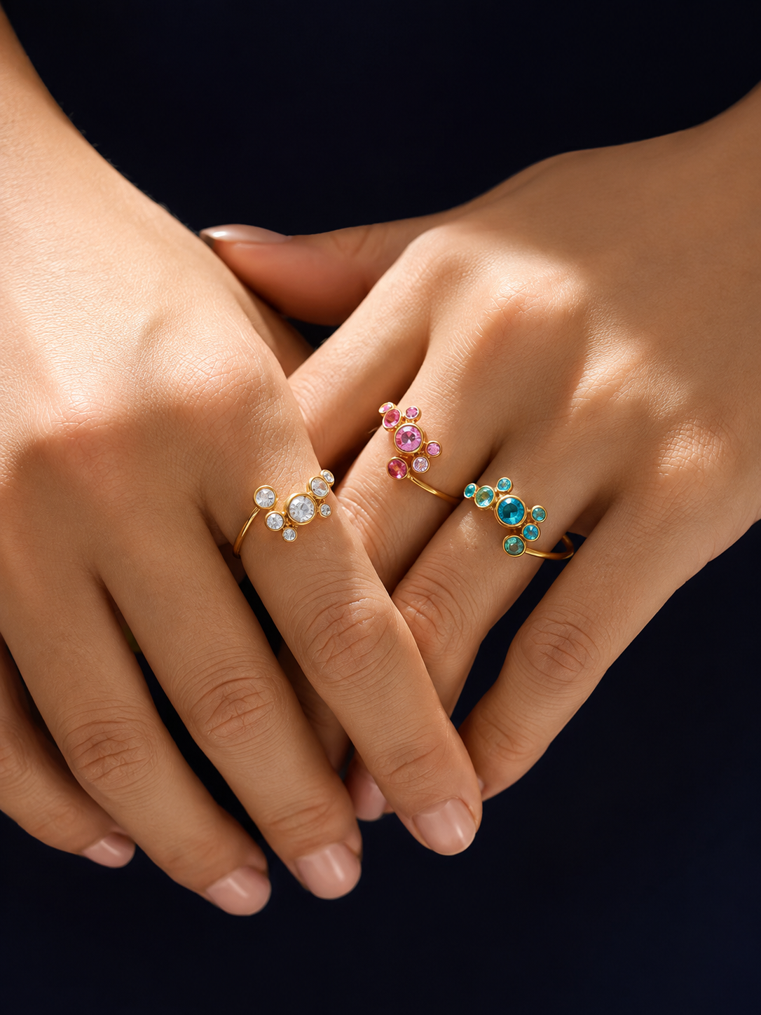 Three delicate gold rings with floral crystal designs are shown on a person's fingers against a dark navy background. The rings feature clear, white, pink, and blue gemstones.