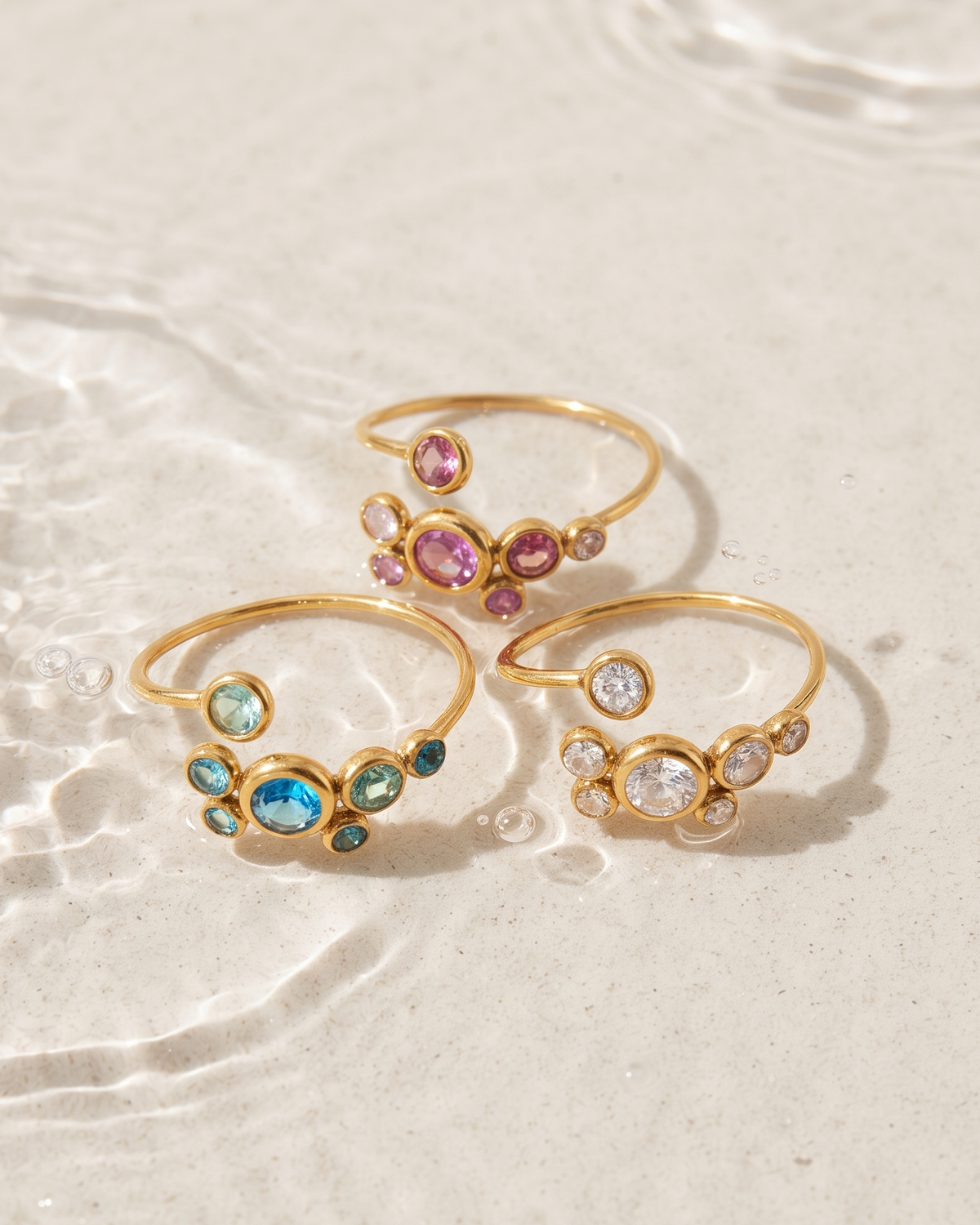 Three gold open-ring rings with colorful gemstones are arranged on a light beige sandy surface with shallow water ripples in the background.