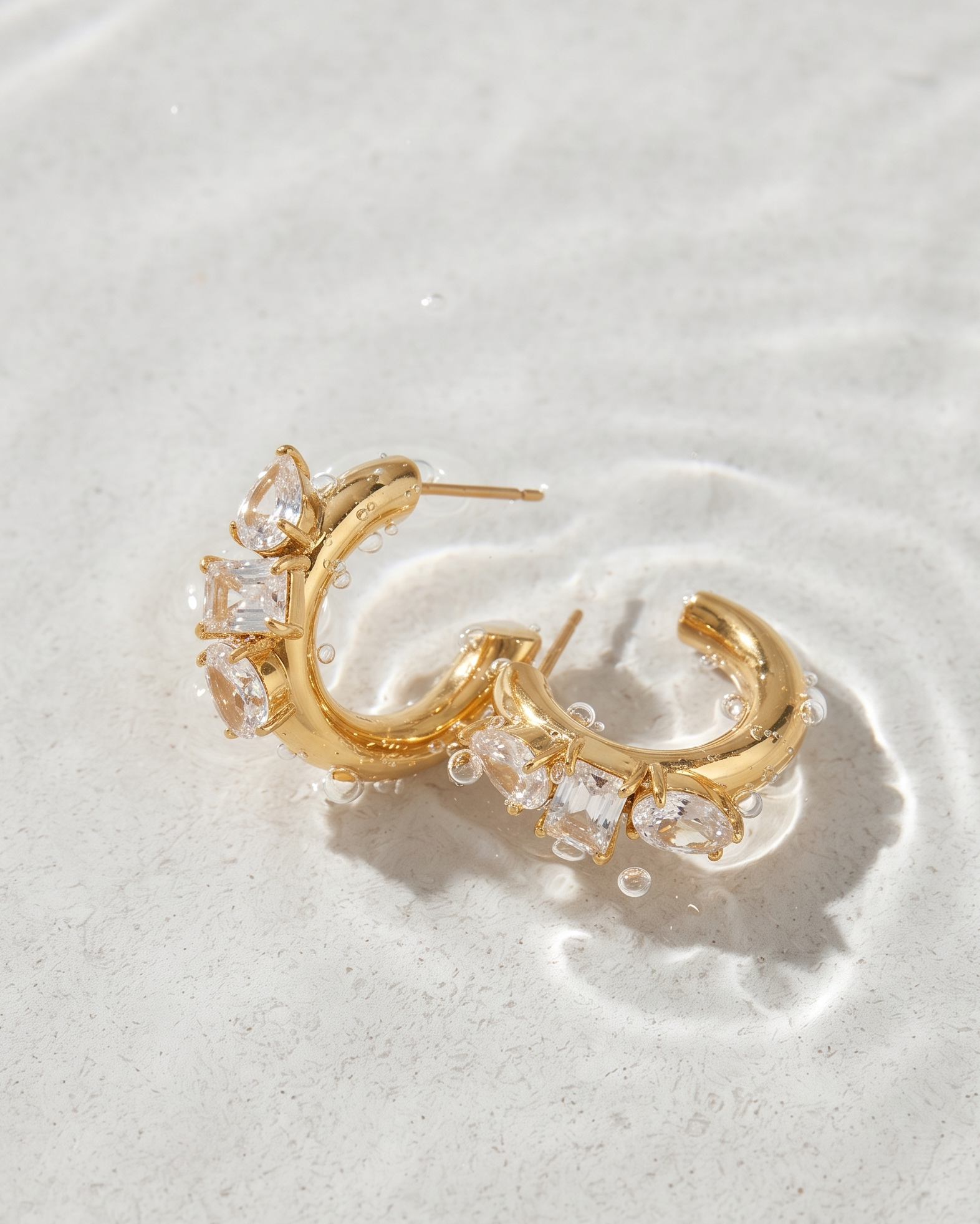 Gold crescent-shaped hoop earrings adorned with clear rhinestones, resting on a textured light gray surface.