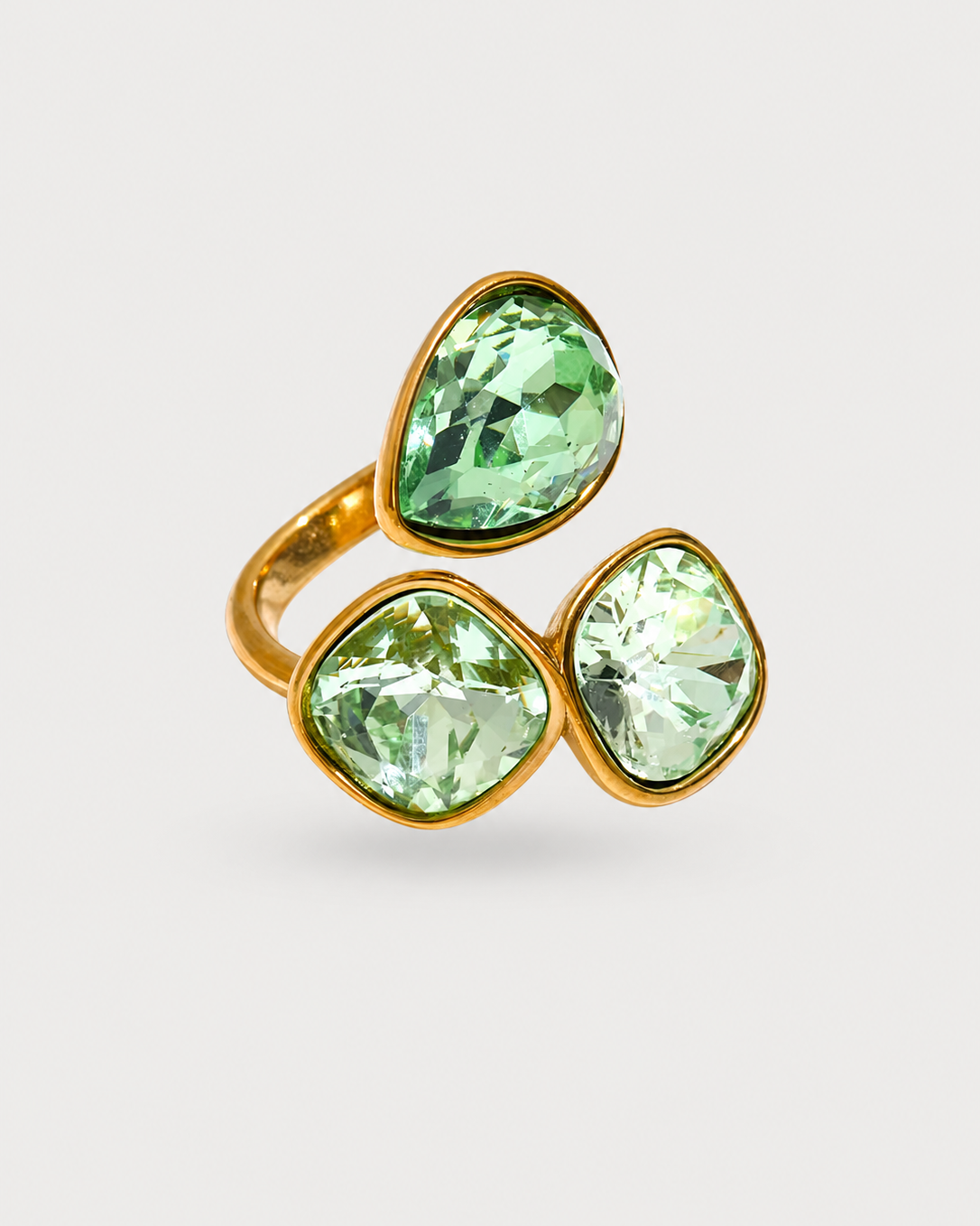 A gold ring featuring three faceted light green gemstones in a triangular arrangement, set against a clean light gray background.