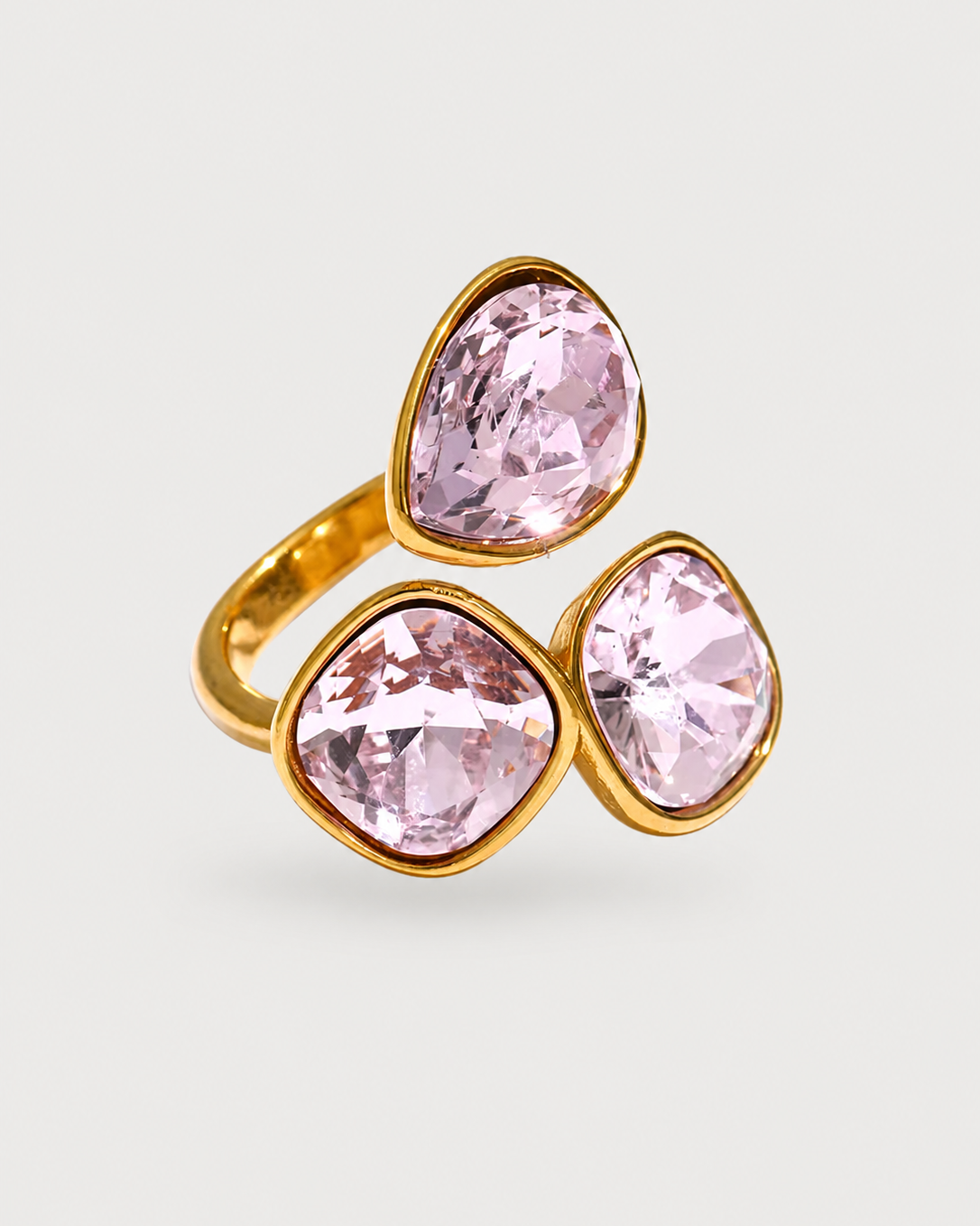 A gold ring featuring three large, faceted light pink gemstones in a triangular cluster setting, shown on a light gray background.