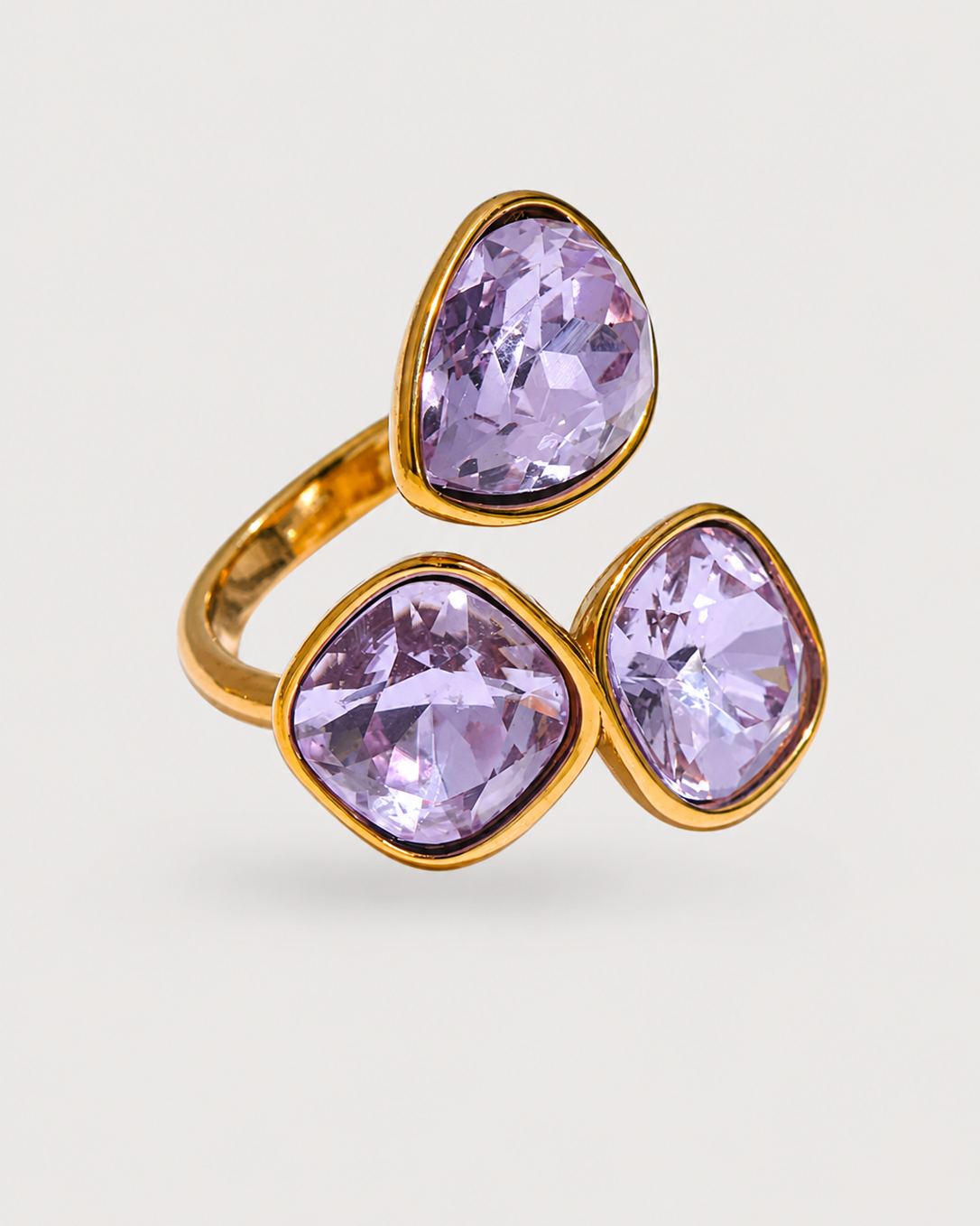 A gold ring featuring three large, faceted lavender-purple gemstones in a cascading triangular arrangement, set against a light gray background.