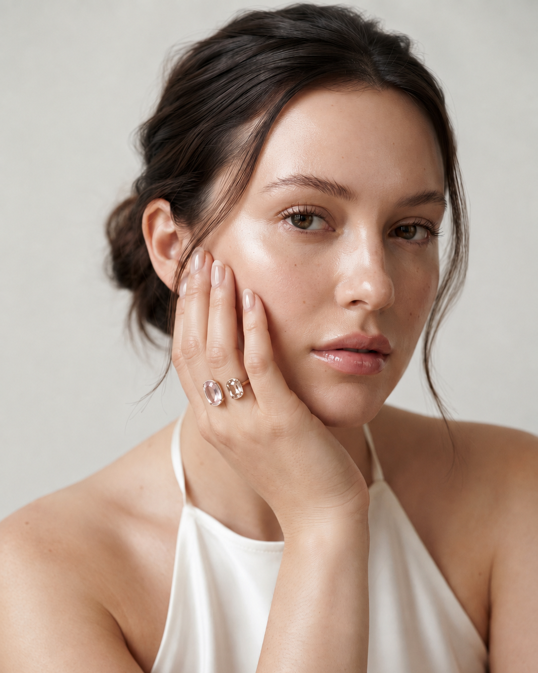A close-up portrait of a woman with her hand resting against her cheek, wearing a delicate ring with two small clear stones in a gold setting. She is wearing a white halter top against a soft, light gray background.