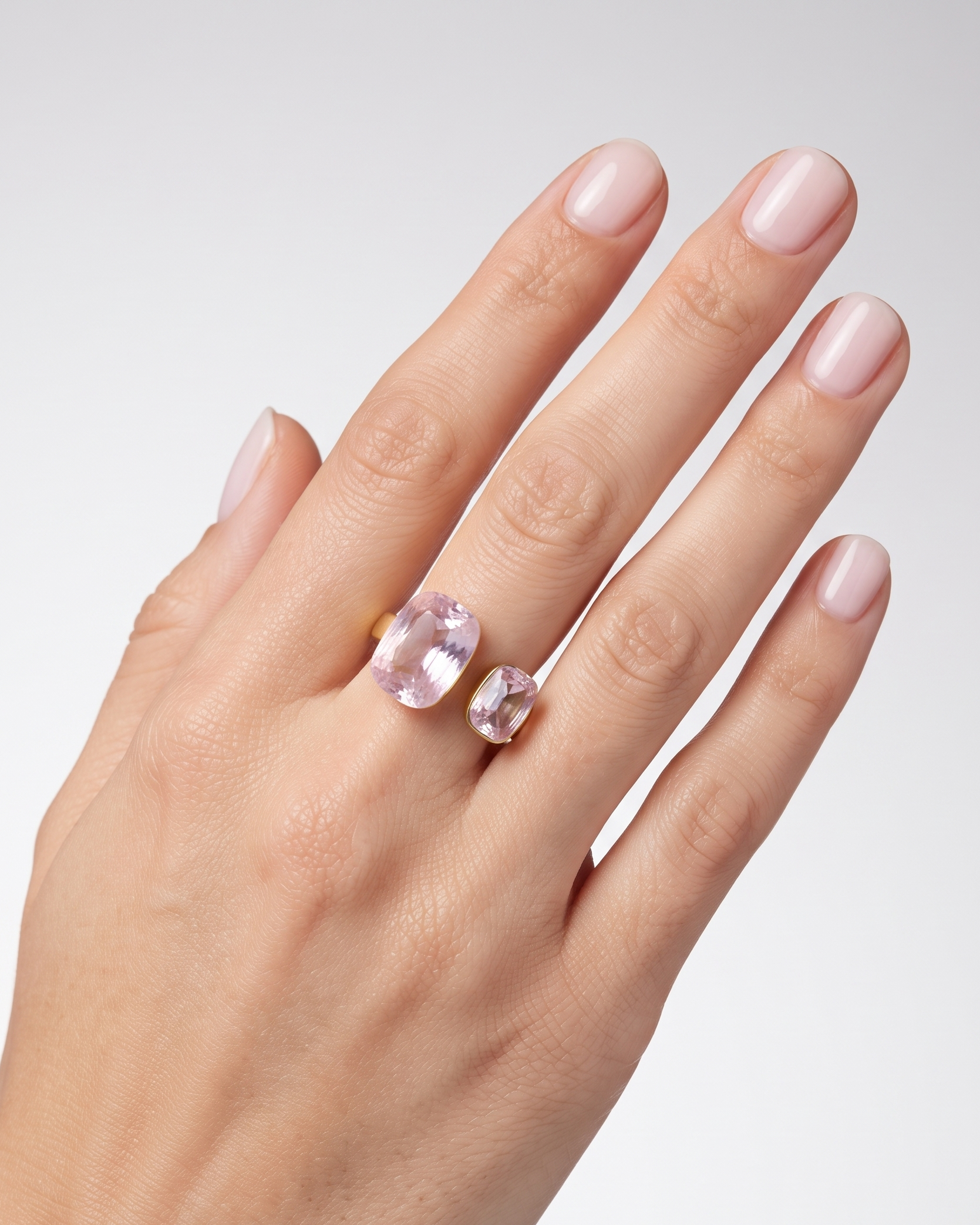 A close-up of a hand wearing two rings: a larger gold ring with a pink oval gemstone and a smaller matching pink gemstone ring on the adjacent finger, set against a light gray background.